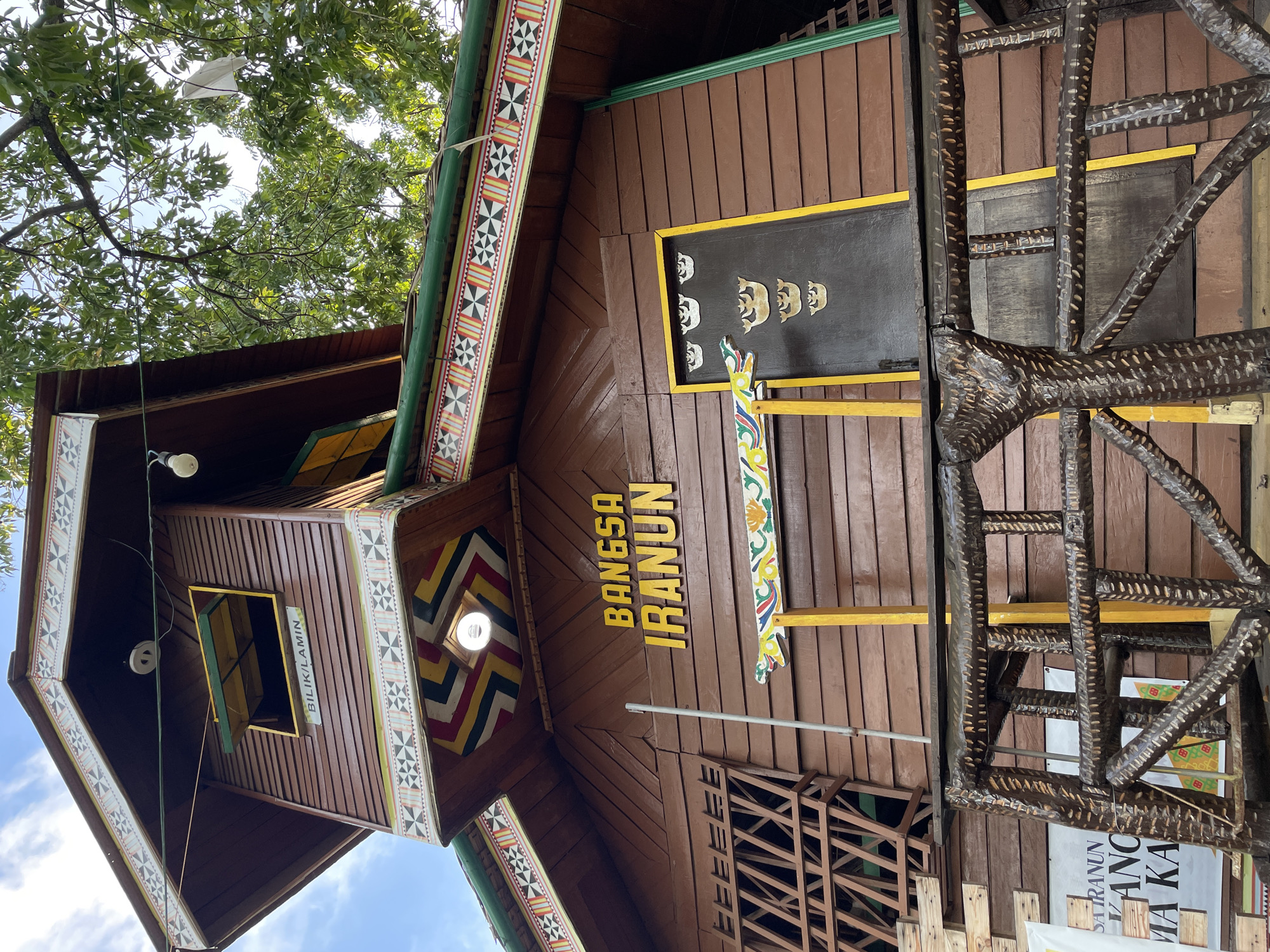 The image shows a traditional wooden building with intricate cultural designs. A prominent sign on the building reads "BANGSA IRANUN" in yellow capital letters. Above the sign, a window is visible with the text "BILIK/LAMIN" on it, and decorative patterns adorn the roofline and the small structure extending from the main building. A dark door with golden crown-like motifs is to the right of the sign. The building's facade is made of horizontal wooden planks. In the foreground, a rustic, carved wooden railing with a textured surface suggests an outdoor viewing area or entrance. A banner with text, partially visible, is mounted on the wall near the railing. The background features lush green foliage from trees and a bright sky. This scene is located in Davao City, Philippines, specifically at the "Bangsa Iranun" exhibit within the Museo Dabawenyo, which showcases the heritage and culture of the Iranun people, an indigenous group in Mindanao.