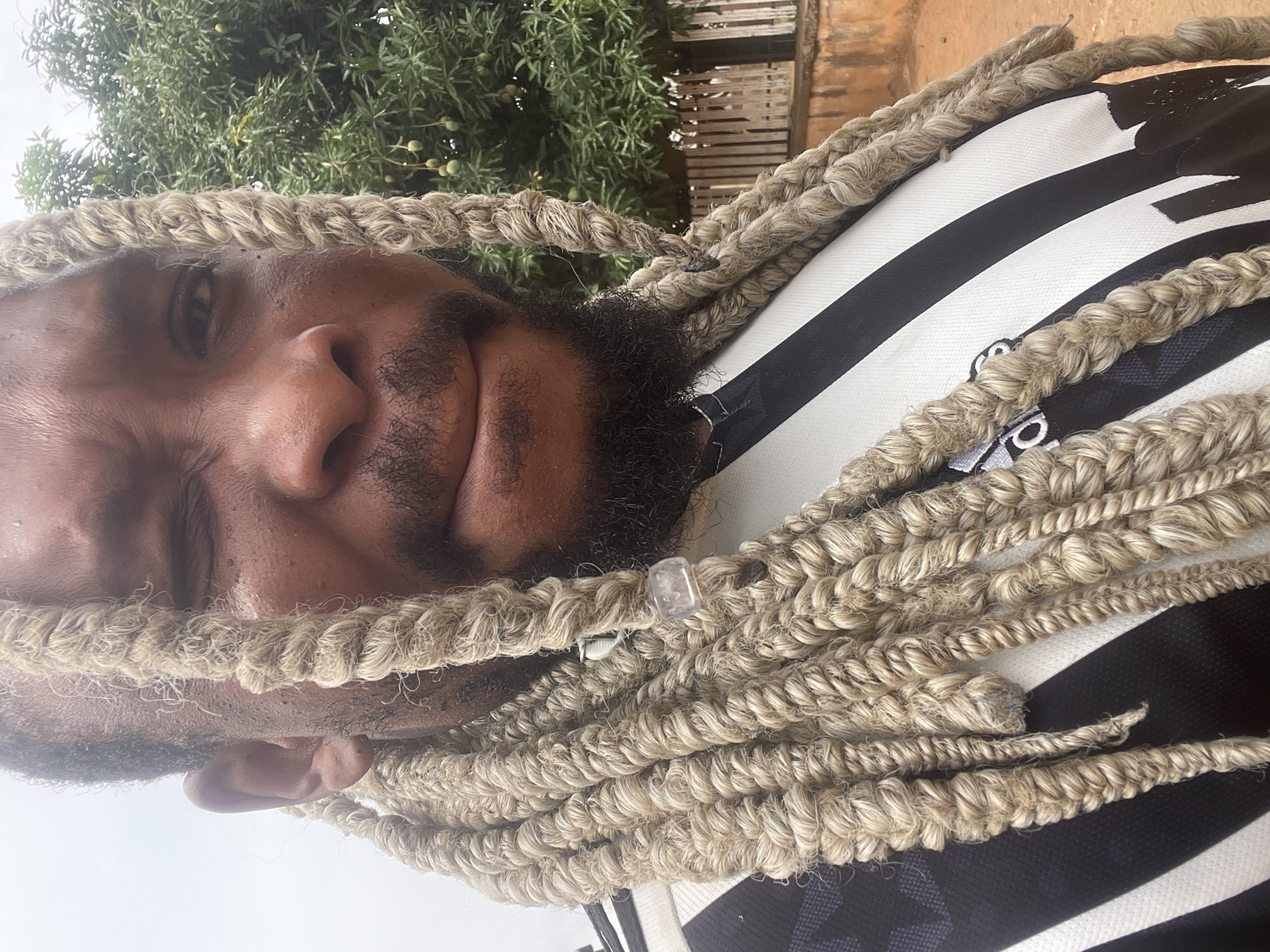 A close-up, tilted shot of a person with light brown dreadlocks. The person has dark skin, a beard, and is wearing a black and white striped shirt. A clear bead is visible in one of the dreadlocks. The background shows green foliage and a building or wall in a muted orange color. The city is Jos, Nigeria.