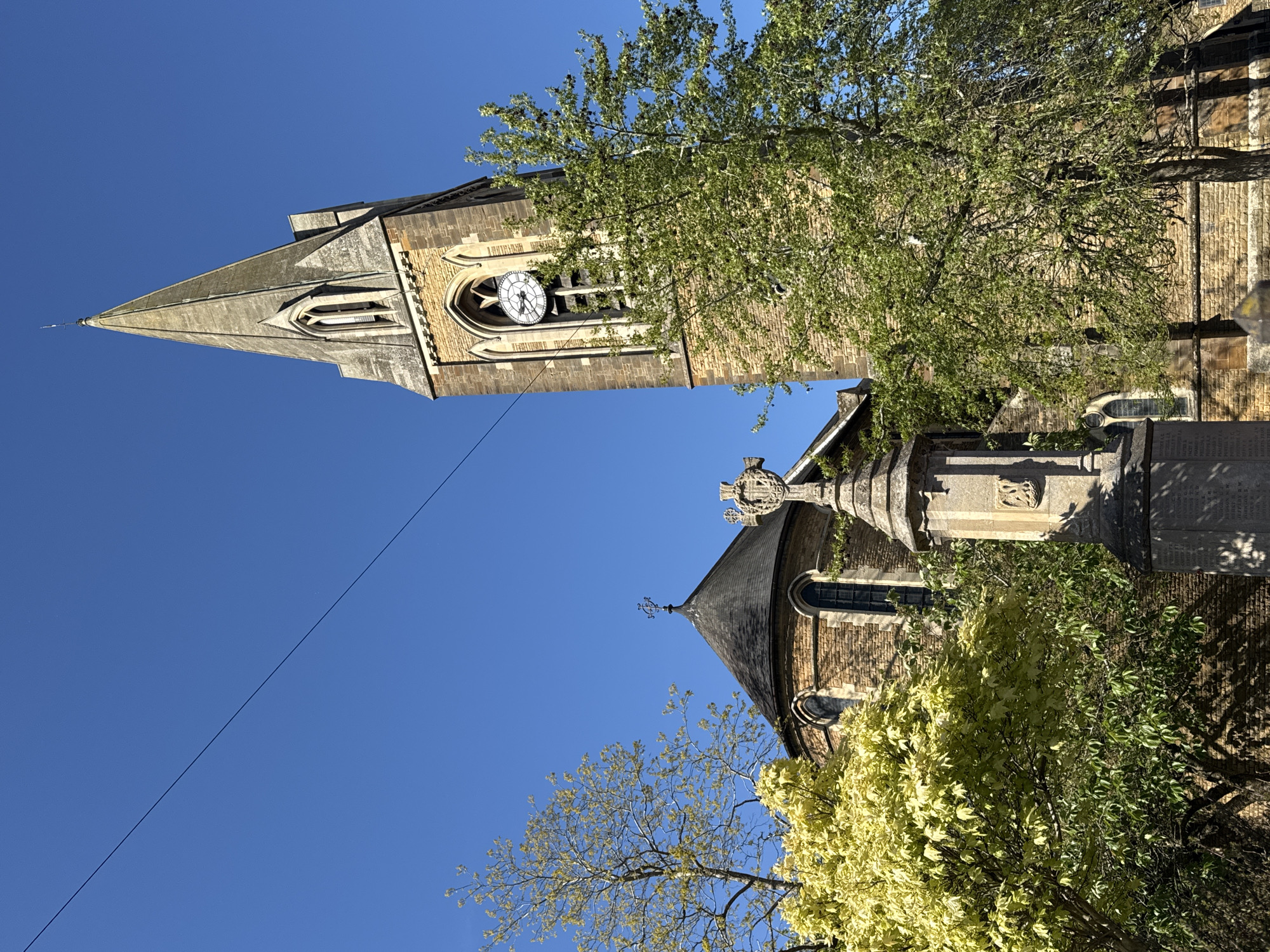 The image captures a historic church and its surroundings in Northampton, United Kingdom, under a clear, bright blue sky. The focal point is the church's tall stone spire and tower, featuring lancet windows and an ornate clock face near the top. The clock's hands indicate a time of approximately 10:10. The tower is built from light-colored stone masonry, showcasing intricate architectural details.
To the right, partially visible behind a tree with lush green leaves, is another section of the church building, appearing to have a rounded apse or chancel with a smaller spire or weather vane at its peak. In the mid-ground, a stone war memorial stands prominently, topped with a detailed carved cross. The memorial's sides bear inscriptions, likely names of those commemorated, though the text is illegible.
Several trees are scattered throughout the scene. One large tree with dense green foliage partially obscures the church tower and part of the main building. Another tree with distinctively lighter, possibly yellow-green or variegated leaves, occupies the lower right foreground. The bright sunlight suggests a clear daytime. A thin, dark wire or power line stretches diagonally across the upper left portion of the sky. The scene conveys a sense of history and tranquility.