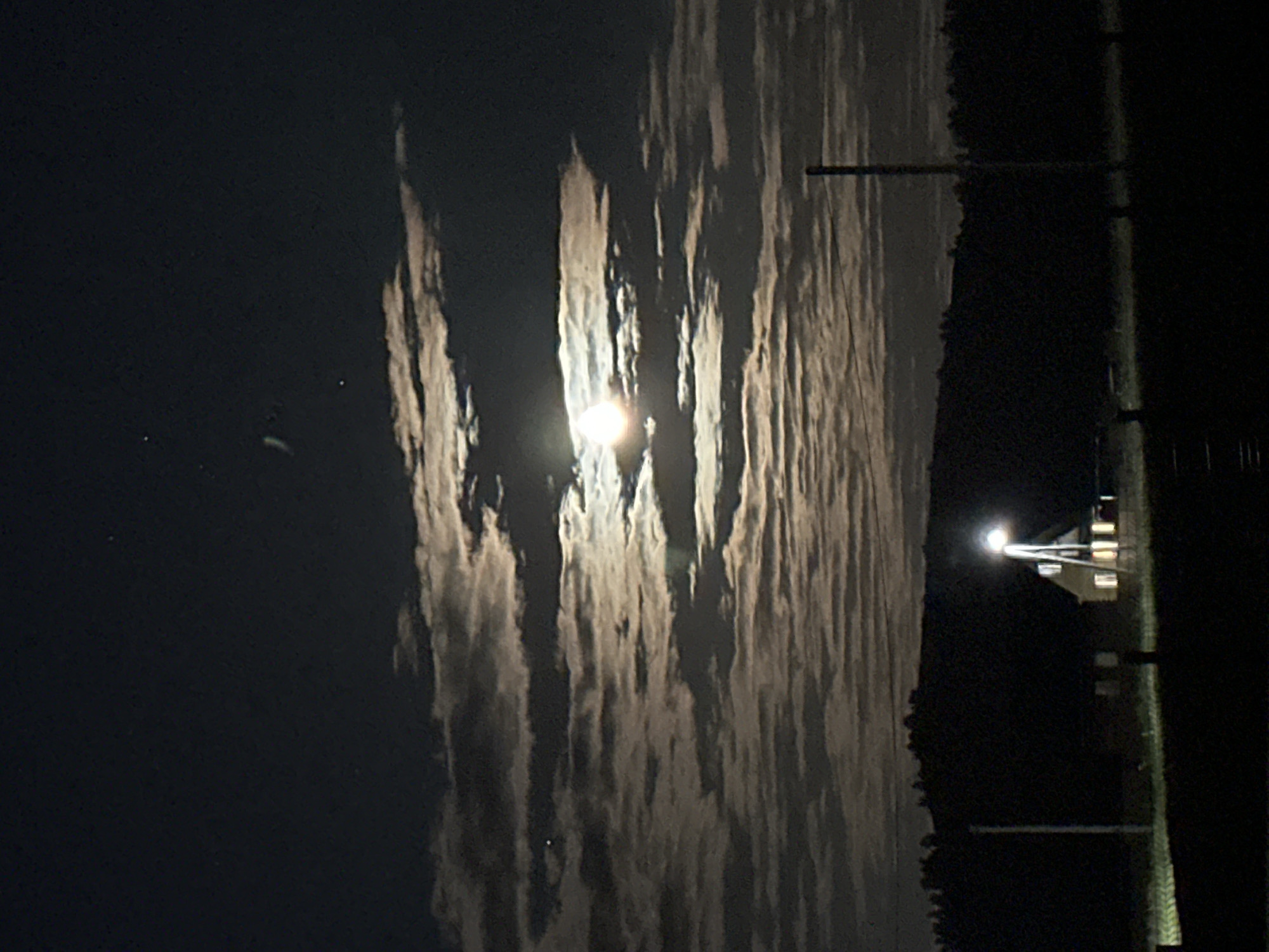 The image depicts a night scene in an unknown location, with a bright full moon dominating the sky, partially obscured by long, striated clouds. A tall utility pole extends vertically on the right side of the image. To the right of the pole is a building illuminated by a bright street light, with a partially obscured roof and windows visible. The foreground is dark, with a suggestion of grass or vegetation.