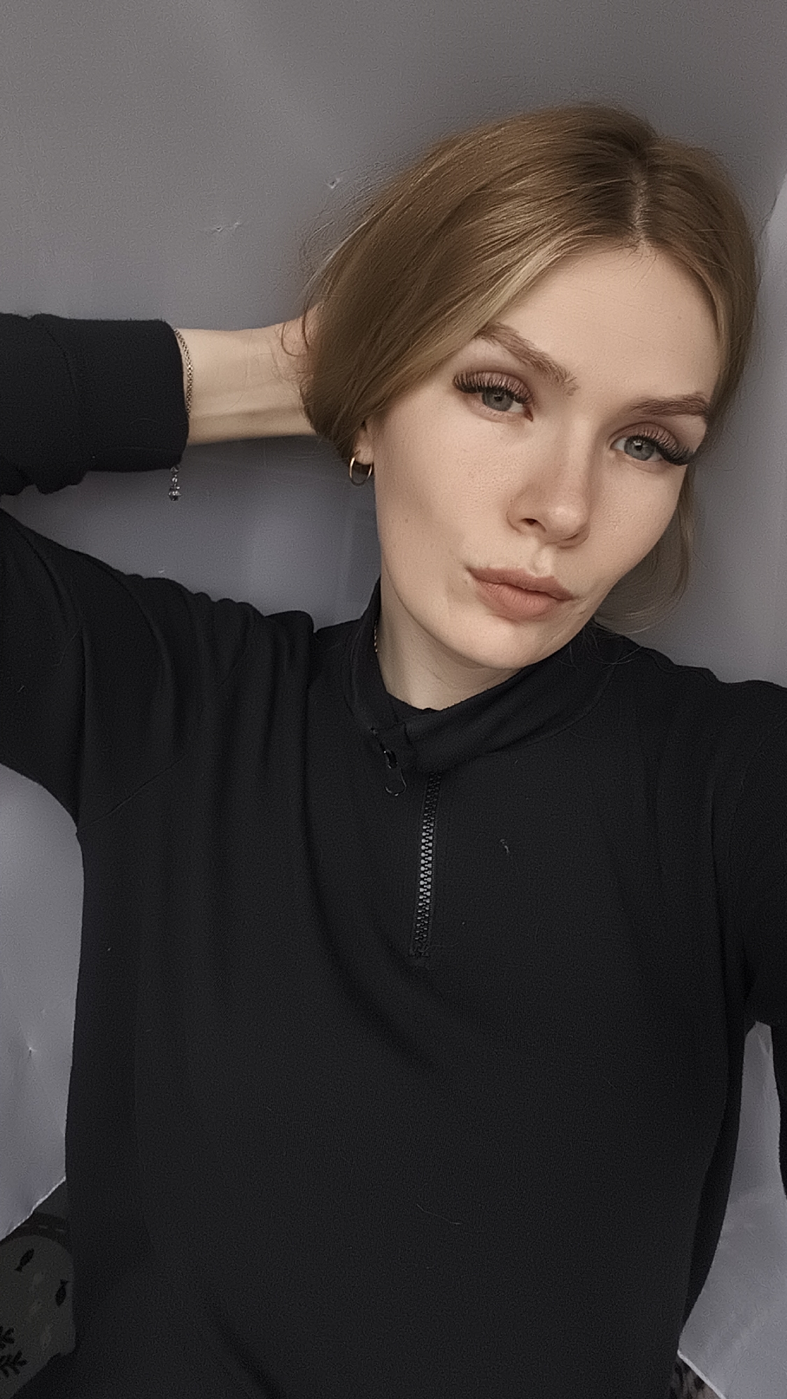 A young woman with light brown hair is posing for a selfie indoors against a gray wall. She wears a black long-sleeved shirt with a zipper and small gold hoop earrings. Her hand rests behind her head, and a bracelet is visible on her wrist. The location appears to be a domestic interior. There is no evidence to connect this image to Русский Вожой, Russia.