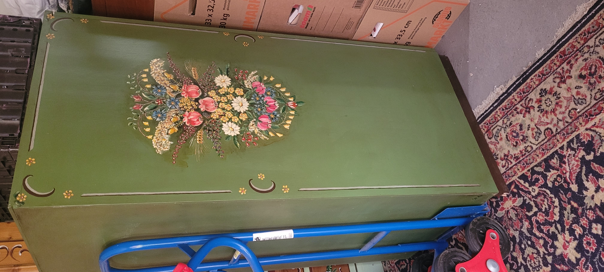 The image shows a tall, olive-green cabinet with a hand-painted floral motif on its front. The floral arrangement is detailed, featuring various types of flowers and foliage in shades of pink, white, yellow, and blue, with some red accents. Decorative borders with crescent moon and flower details are painted on the cabinet's edges. To the left of the cabinet, a blue and red dolly is positioned, with its wheels visible on a patterned rug. In the background, several cardboard boxes are stacked, with text indicating they are likely for moving or storage, and some text is in German. The environment appears to be indoors, possibly a storage area or a room undergoing renovation or packing, given the presence of the dolly and boxes. The overall impression is one of an indoor space with stored items.