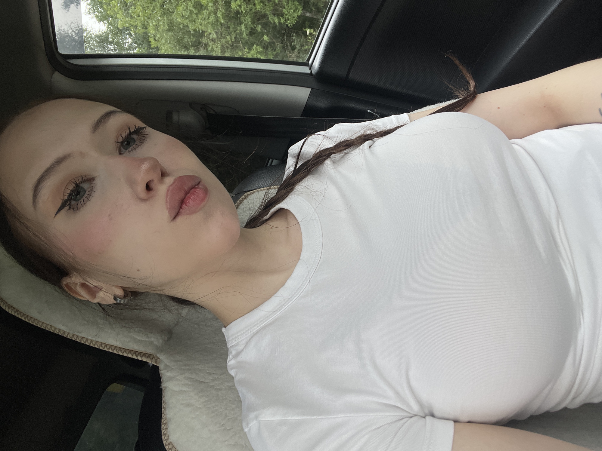 A young Caucasian woman is photographed from the chest up, seated in the passenger seat of a vehicle. She has fair skin, light-colored eyes, and dark brown hair styled in a braid that falls over her shoulder. Her makeup includes winged eyeliner, mascara, and pink blush. She is wearing a plain white t-shirt. Her lips are slightly parted, giving the impression she is about to speak or has just spoken. The background shows the interior of a car, including the car seat and a side window with greenery visible outside, suggesting a rural or suburban setting. There are no specific landmarks or elements in the image that can definitively place the scene in Gavrilovka, Russia. The lighting is natural, indicating it's daytime.