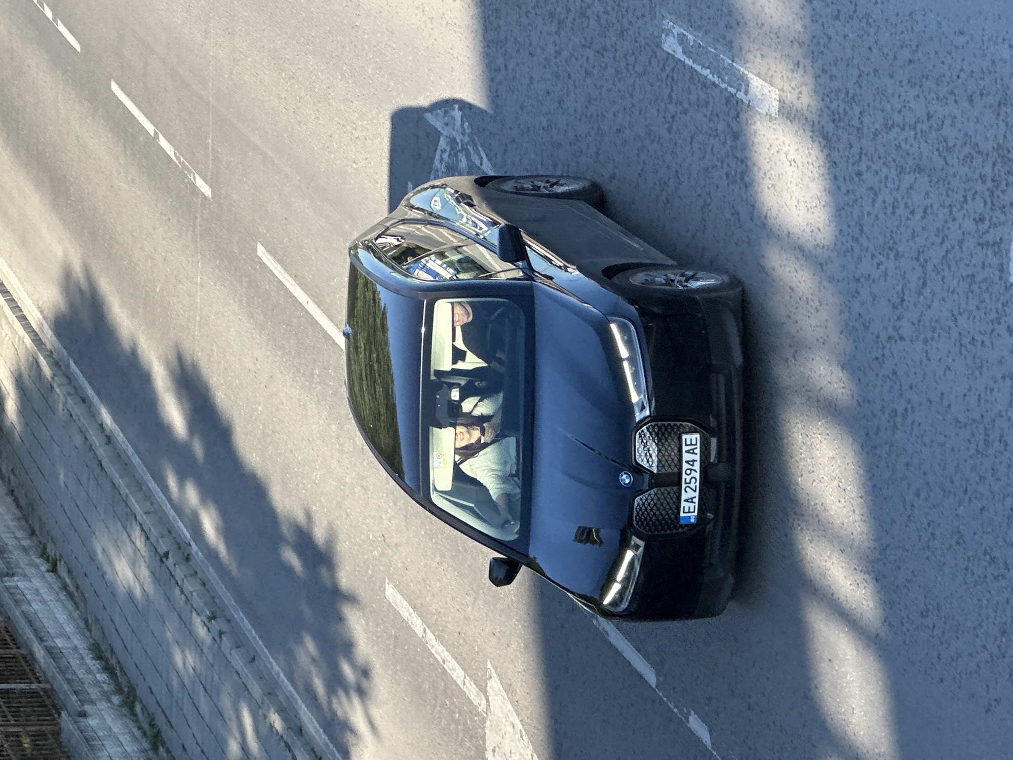 A black BMW iX car, bearing the license plate EA2594AE, is travelling along a road marked with white lane dividers. Two individuals are visible inside the car. The scene appears to be Veliko Tarnovo, Bulgaria, based on the license plate. The picture appears to have been taken from an elevated perspective, looking down at the car. Shadows suggest sunlight is present. A concrete barrier lines the edge of the road.