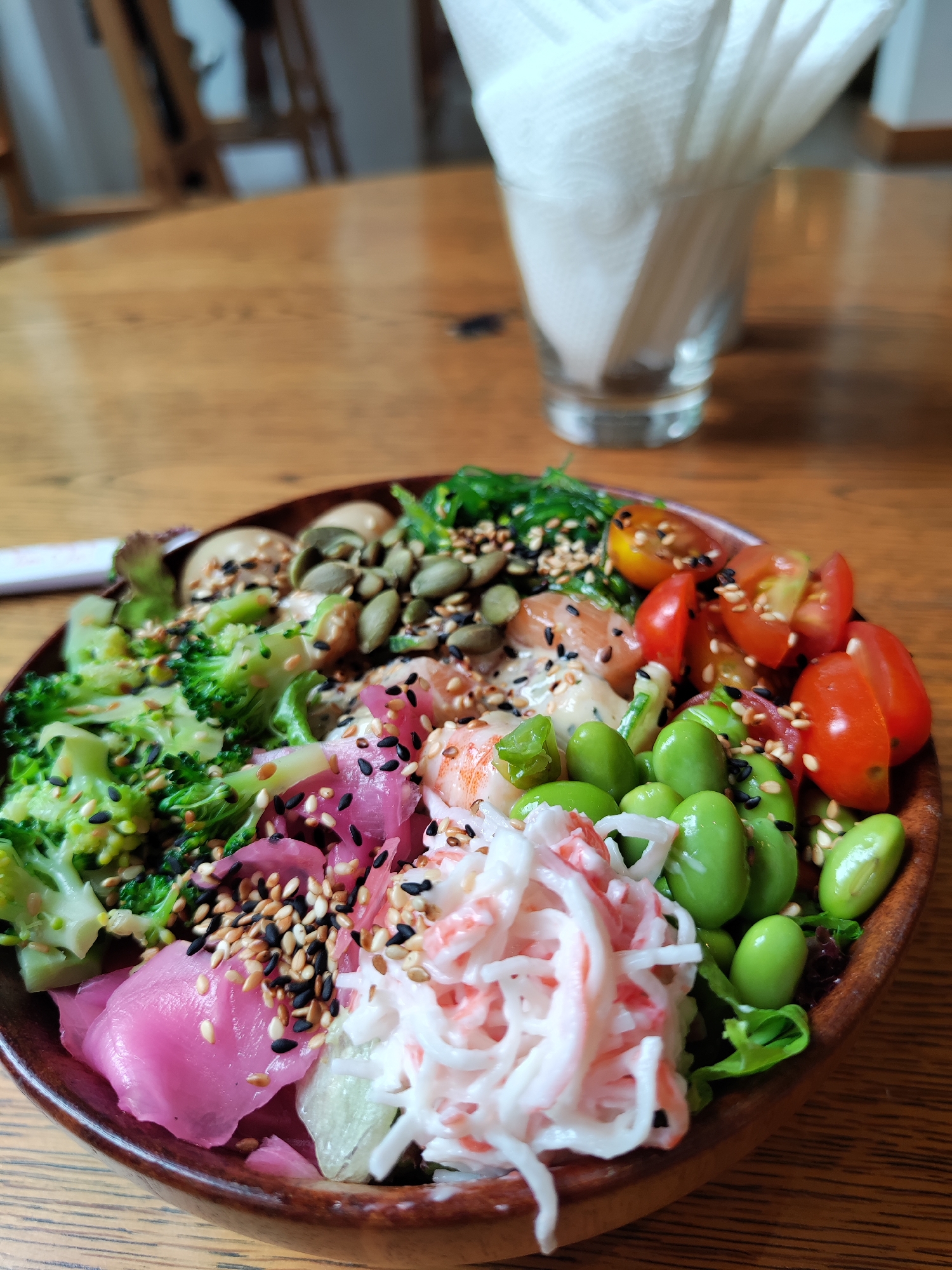 A close-up shot shows a poke bowl on a wooden table in a restaurant or cafe. The bowl contains ingredients such as broccoli, edamame, imitation crab meat, seaweed salad, cherry tomatoes, pickled ginger, and other items, possibly including various seeds and sauces. A glass filled with napkins is visible in the background. The location is in Thủ Đức, Vietnam.