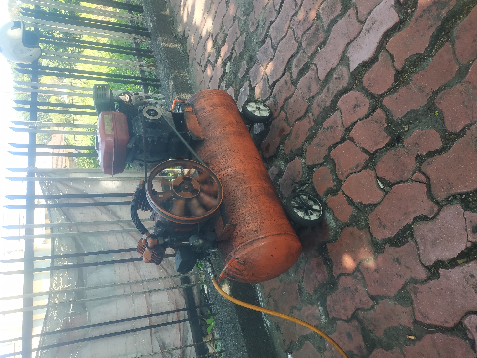 The image shows a red air compressor with a black engine and a pulley system. It is situated on a paved walkway made of red interlocking bricks. The air compressor is equipped with two small wheels and a yellow hose is attached to it. In the background, there is a black metal fence with vertical bars, behind which a white tarp is hung. Beyond the fence, trees and foliage are visible. A white helmet is hanging on top of the fence. The scene appears to be outdoors, likely in a residential or commercial area in the city of Jember, Indonesia, given the common presence of such fences and paved walkways in the region. The lighting suggests it is daytime, with sunlight casting shadows on the bricks. No people are present in the image.