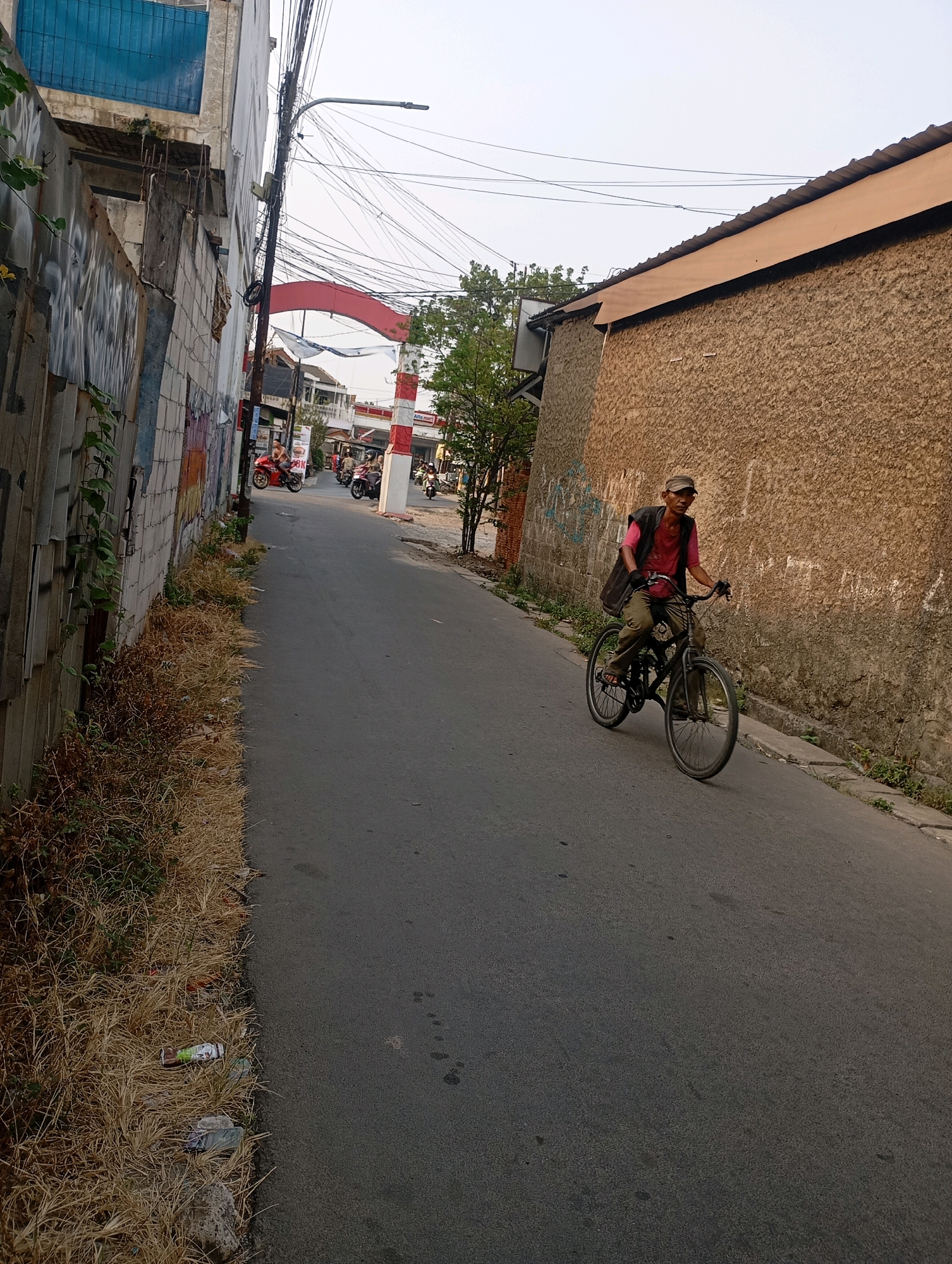 In Babelan, Indonesia, a man rides a bicycle down a narrow street. The man is wearing a pink shirt, a vest, and a cap. He is carrying a bag on the back of his bicycle. The street is lined with buildings on both sides. On the left, there is a tall building with a corrugated metal fence and graffiti. On the right, there is a rough-textured building with a brown, stucco-like exterior. The sky is overcast. In the background, there is a red archway and several motorbikes are parked. There are also power lines strung across the sky. The ground is asphalt, and there is some dry grass and debris on the side of the road.