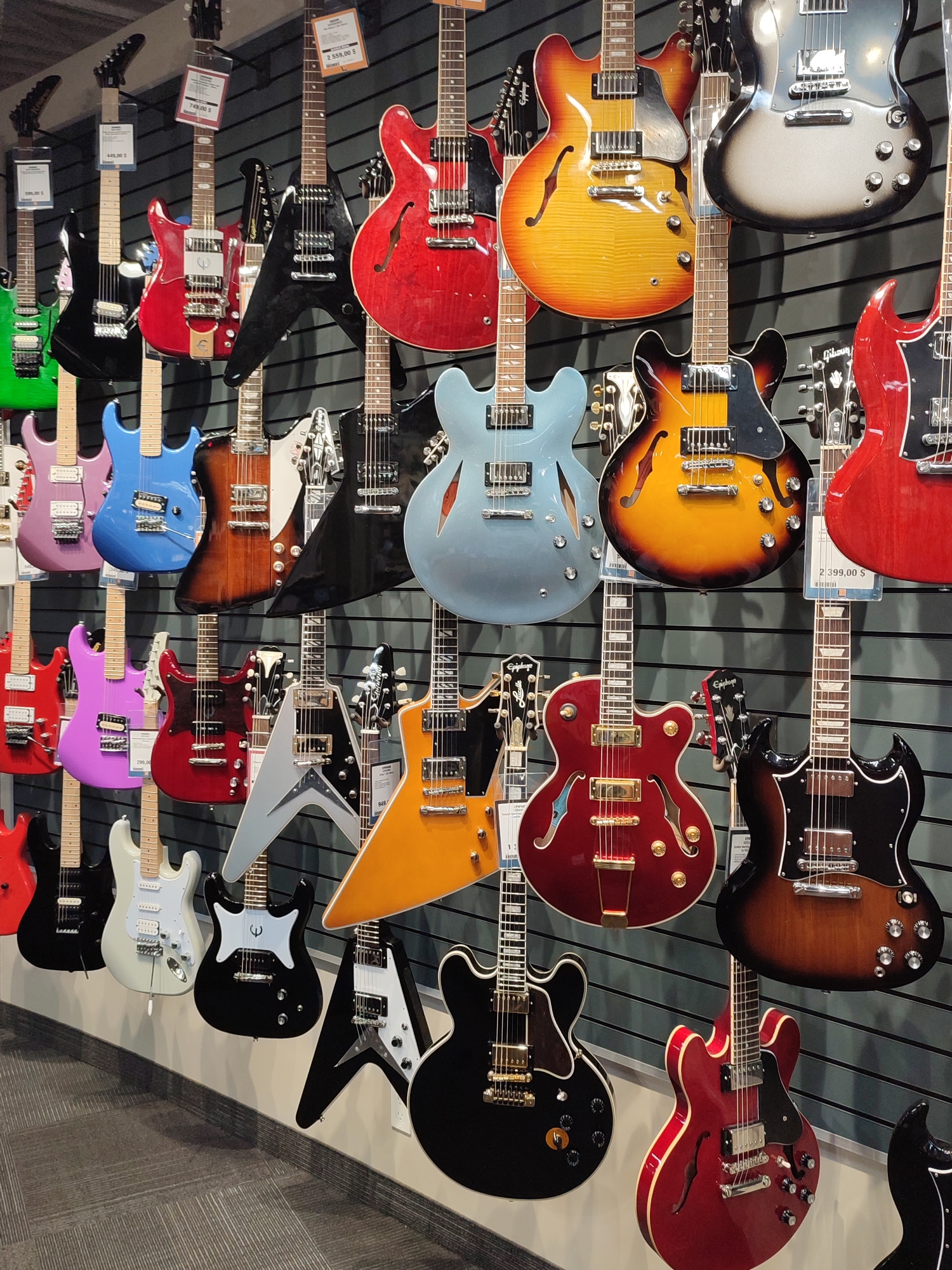 This image captures a display of electric guitars in a retail store, possibly a music shop, located in Longueuil, Canada. The guitars are hung vertically on a dark gray, slatted wall. The guitars vary in color, shape, and style, with prominent examples including red, blue, green, black, and sunburst finishes. Several guitars have distinct designs, such as sharp angles and cutaways. Price tags are visible on some of the instruments. The foreground shows a gray carpeted floor and a white baseboard along the wall. No people are visible in the scene.