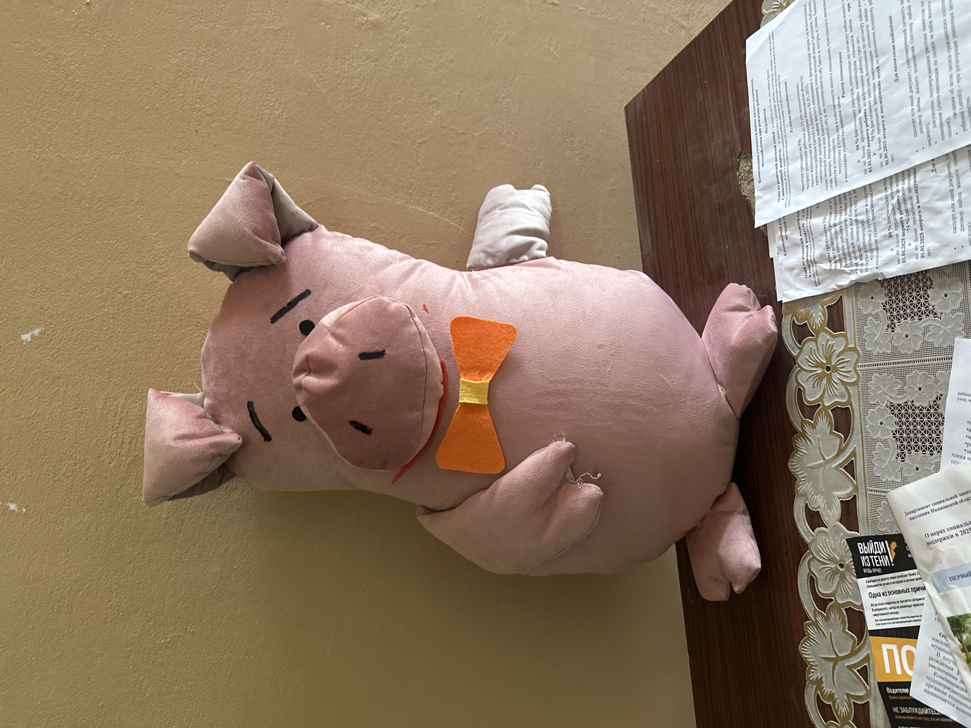 A plush toy pig, pink with a simple face and an orange bow tie, sits on a wooden surface. The pig is positioned centrally and faces forward, its ears tilted slightly to the sides. To the right of the pig, a portion of its arm is visible. The wooden surface is cluttered with several white documents, some partially obscured by the pig and a decorative lace runner. The background consists of a textured, pale yellow wall. The overall scene appears to be indoors, possibly in a room within the city of Gavrilov Posad, Russia, given the context of the image.