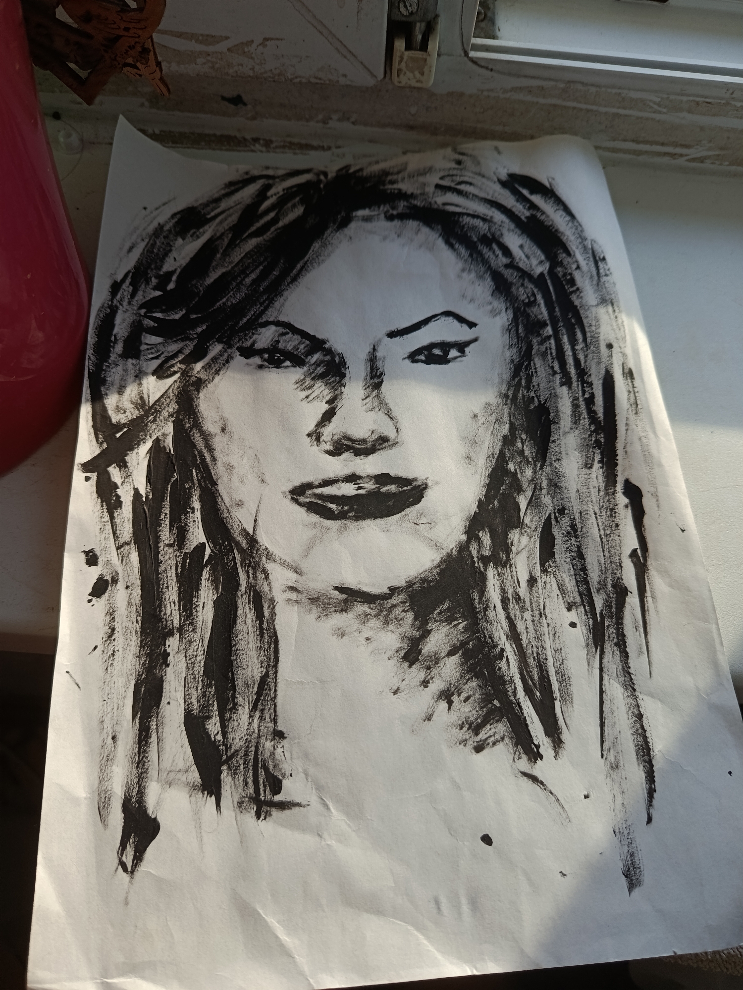 A charcoal drawing of a woman's face is lying on a surface, possibly a windowsill. The drawing features expressive, thick brushstrokes forming the woman's hair, face, and neck. The artwork has a somewhat unfinished quality. The background shows parts of a window frame. The location cannot be determined to be Moscow, Russia from the visual information provided.