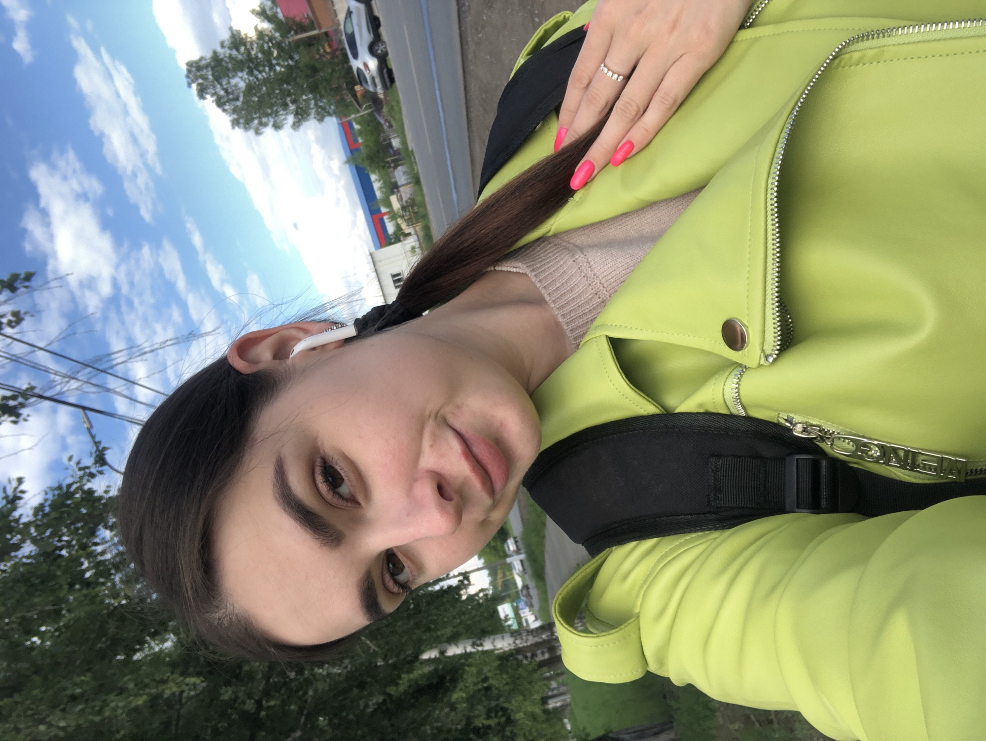 The image is a tilted, close-up selfie of a young woman taken outdoors in Chaikovsky, Russia, during daytime. She has dark hair, dark eyebrows, and brown eyes, looking towards the camera with a calm, soft expression.
She is wearing a vibrant lime green jacket, possibly made of faux leather, featuring prominent silver zippers. A beige ribbed knit top is visible underneath the jacket. Her right hand, adorned with bright pink nail polish and a silver-toned ring with multiple small stones on her ring finger, rests on her left shoulder. A white earbud is visible in her left ear, and a black backpack strap crosses her left shoulder. The zipper pull on her jacket clearly displays the engraved text "MEIN GO".
The background features a bright blue sky with scattered white clouds. Green trees with lush foliage are visible, suggesting a natural outdoor setting. Faintly in the distance, a road, a white car, and part of a building with blue and red sections can be seen, indicating an urban or suburban environment.
