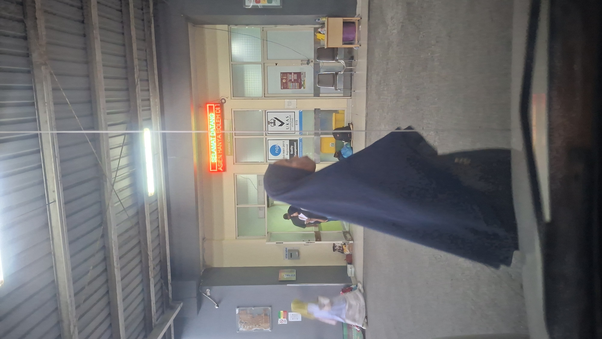 The image depicts the entrance of a building, possibly a hospital or clinic, with a neon sign that reads "SELAMAT DATANG" (Welcome) and "ASIEN HANYA BOLEH DII" (Patients are only allowed in). The scene is set in the city of Banda Aceh, Indonesia.
In the foreground, a person wearing a dark blue hijab is seen from the side, walking. To the left, another person in a light-colored hijab and dress is seated on the ground, holding a white container. Further back, two individuals are visible through an open doorway. One is wearing a dark shirt and the other a lighter colored shirt.
The environment appears to be an indoor-outdoor space, with a covered walkway leading to the building's entrance. The walls are painted in muted colors, and there are several signs and posters displayed. There are also chairs, a small table with a purple bin, and bags scattered around. The lighting suggests it might be evening or indoors with artificial lights. The overall impression is one of a functional public space, possibly with some waiting areas.