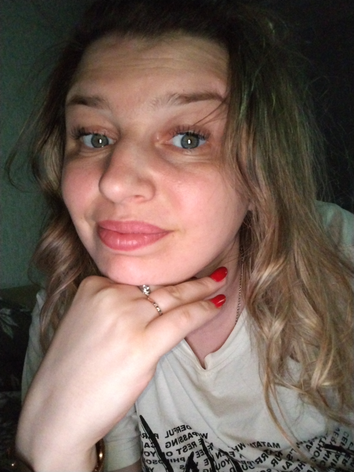 A close-up selfie of a woman with wavy blonde hair and green eyes, with her hand resting on her chin. She has red nail polish and is wearing rings. The background is dark, and she is wearing a white t-shirt with printed words. The location is Unknown, Unknown.