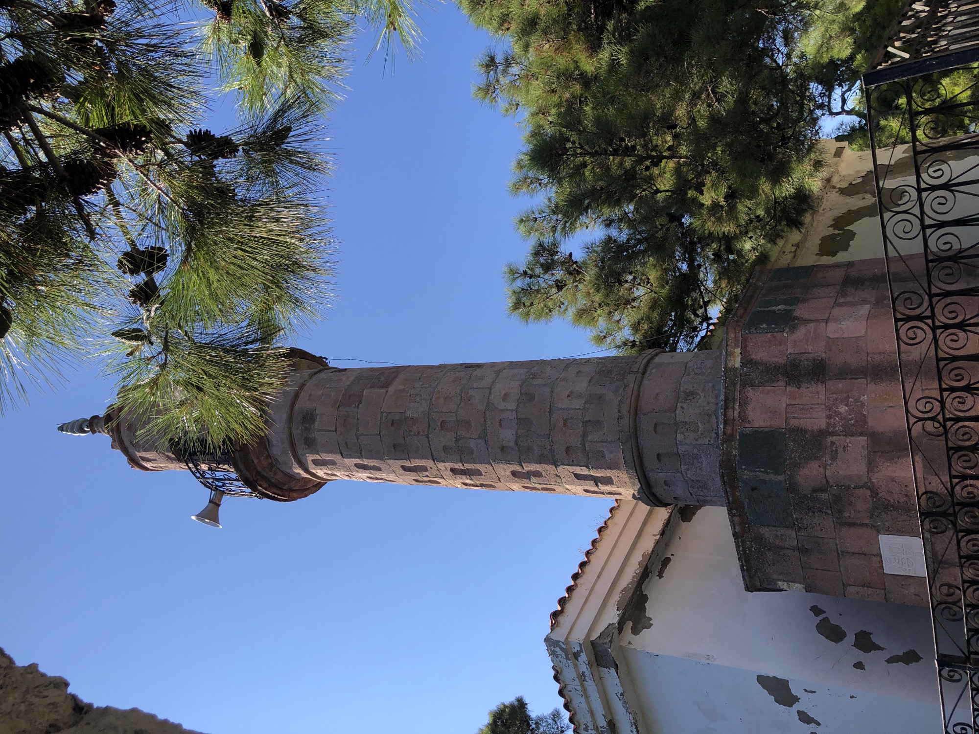 A tall, cylindrical stone minaret stands adjacent to a white building with a tiled roof in Karaburun, Turkey. The minaret is constructed of reddish-brown stones and has a decorative balcony with a speaker. Green pine trees with cones overhang the minaret and a section of the white building. The sky is a clear, bright blue. A black wrought-iron fence is visible in the foreground.