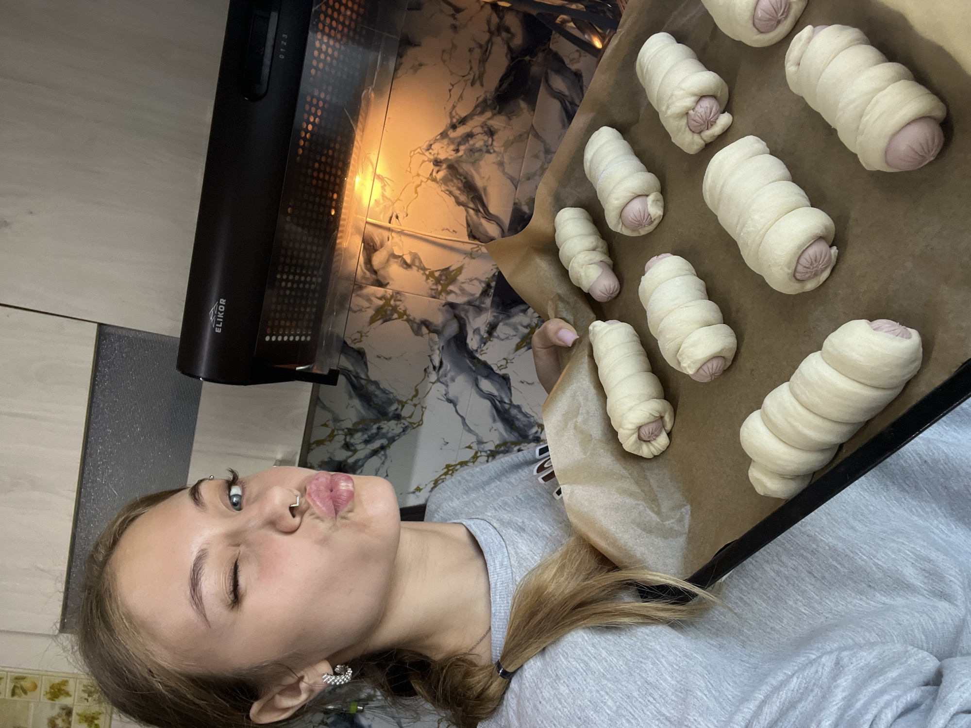 A woman with light skin, light brown hair in a ponytail, and facial piercings is holding a baking sheet with nine raw pigs in a blanket in front of an open oven. The oven is built into a kitchen cabinet. The kitchen's backsplash appears to be marble. The brand "ELIKOR" is visible on the oven hood. The location is Фоки, Russia.