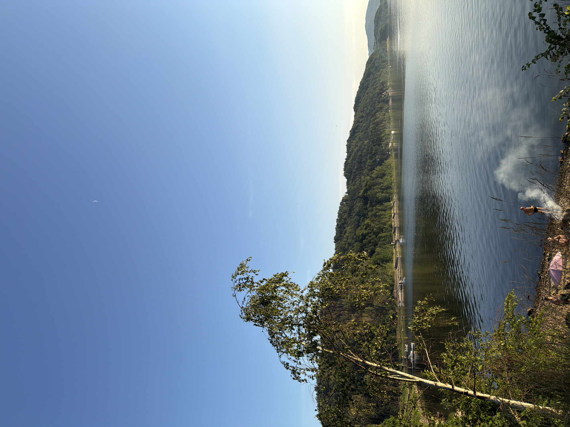 The image depicts a serene lakeside scene in Lipie, Poland. In the foreground, a slender birch tree with delicate leaves leans towards the tranquil water. The shoreline is a mixture of sand and pebbles, where a few individuals are visible. One person stands near a small fire producing wisps of smoke, while another sits beside a pink umbrella. The lake stretches out, reflecting the clear blue sky and the lush green hills that form the backdrop. Several boats are anchored in the distance, adding to the peaceful atmosphere. A faint crescent moon is visible in the upper right corner of the sky.