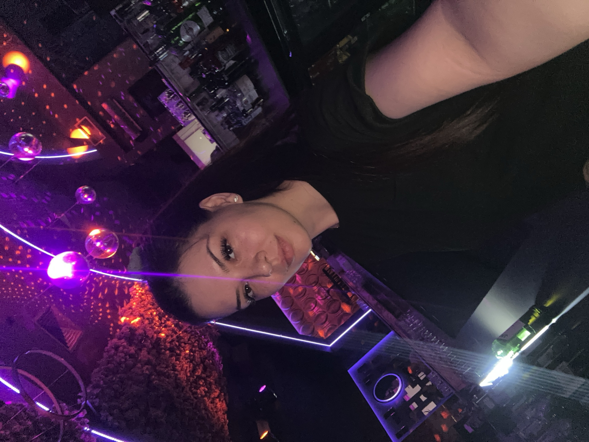 In an indoor setting, possibly a nightclub or bar, a young woman with dark hair and fair skin, wearing a black t-shirt, is taking a selfie. She has makeup on, including eyeliner and lipstick, and is wearing a small earring. The background is dimly lit and features a bar area with bottles on shelves. Above, there are purple neon lights and disco balls, creating a vibrant, colorful atmosphere. The overall impression is a casual, social moment captured in a lively entertainment venue. The provided information about the city of Sliven, Bulgaria, is not directly inferable from the visual content of the image itself.
