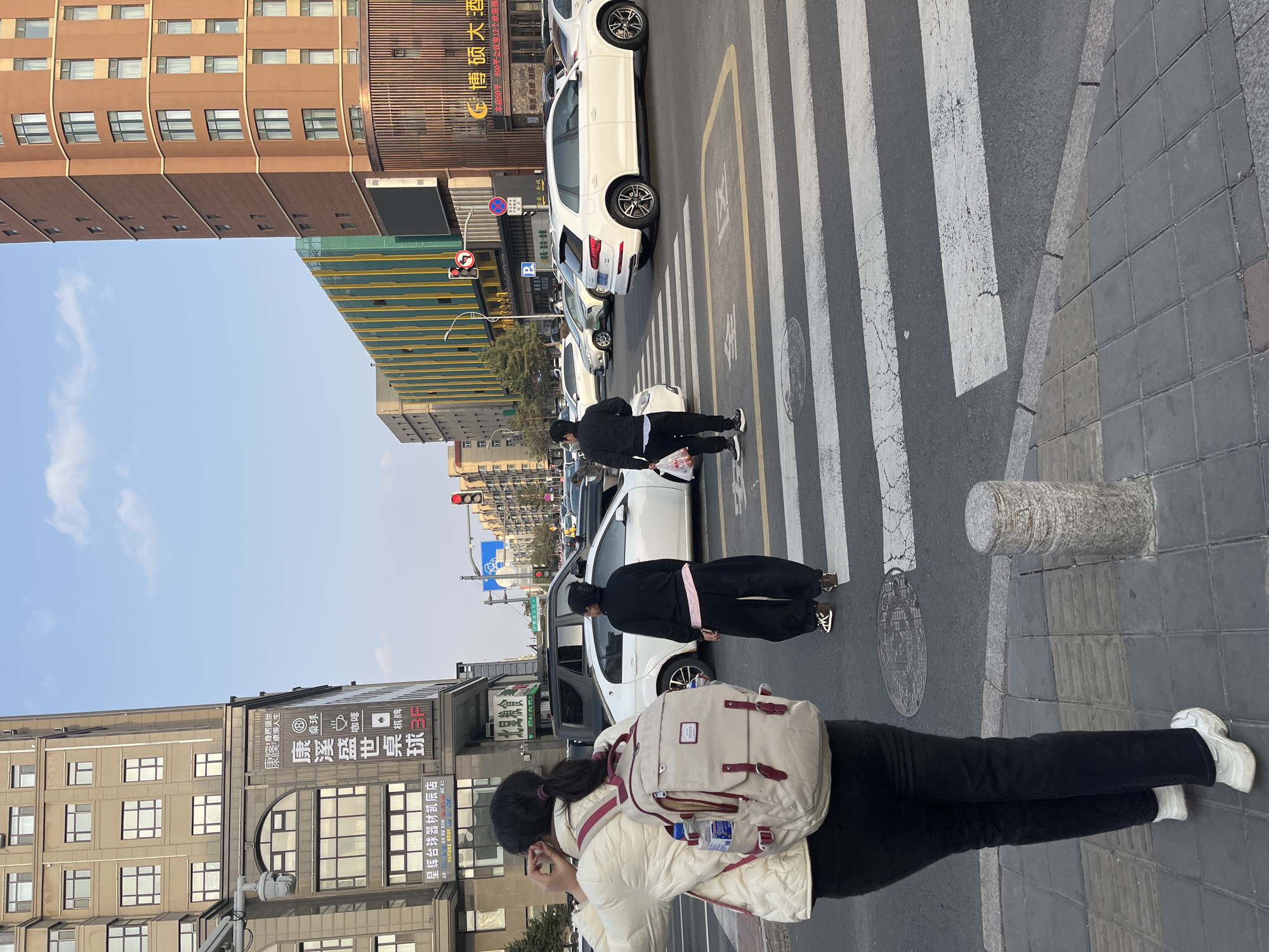 An urban street scene in Changchun City, China, is depicted from a slightly elevated perspective, showcasing a crosswalk, pedestrians, and surrounding buildings under a clear blue sky. In the foreground, a person with dark hair, wearing a white padded jacket and black pants, carries a light-colored backpack with maroon accents and is looking down at a mobile phone. Ahead of them, another individual in a black jacket and black pants stands near a white SUV on the crosswalk. Further down, another person dressed in black is visible.
Several white cars are present on the street, some appearing to be parked or waiting at the crosswalk. The street features a prominent zebra crossing with some visible wear and cracks, as well as a yellow box road marking. To the right, a gray tiled sidewalk with a cylindrical stone bollard separates the pedestrian area from the road.
The background is dominated by multi-story buildings. On the left, a reddish-brown building with numerous windows rises, while another building features a large sign with Chinese characters. Visible text on signs includes "康溪盛世桌球" (Kangxi Shengshi Billiards), with icons for "桌球" (Billiards), "咖啡" (Coffee), and "棋牌" (Chess and Cards). Other promotional text reads "身居盛世 康溪人生" (Living in a flourishing age, Kangxi life) and "本店60平-800平会议室12个欢迎预" (Our store has 12 meeting rooms from 60sqm-800sqm, welcome to reserve). Another sign mentions "博硕大酒" (Boshuo Great Wine/Hotel) and "星辉台球器材凯辰店" (Xinghui Billiards Equipment Kaichen Store). A traffic sign with a red circle and a slashed left arrow indicates a "No Left Turn" restriction. The scene captures a typical bustling daytime street environment.