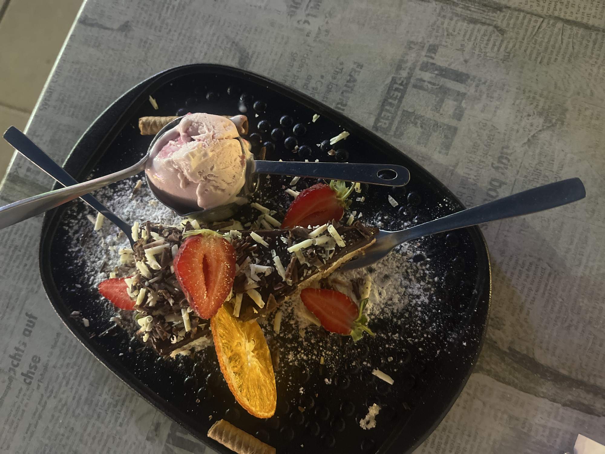 A close-up, overhead shot features a dessert on a black, oval-shaped plate. The dessert appears to be a slice of chocolate cake topped with chocolate shavings, white chocolate shavings, sliced strawberries, and an orange slice. A scoop of pink ice cream sits on a spoon resting on the dessert. Two wafer rolls are also present on the plate. The plate is placed on a surface covered with what appears to be newspaper or a similar patterned material. The lighting suggests an indoor setting, possibly a restaurant or cafe. The image does not provide any specific details that would allow for the identification of its location in the city of Nesebar, Bulgaria.