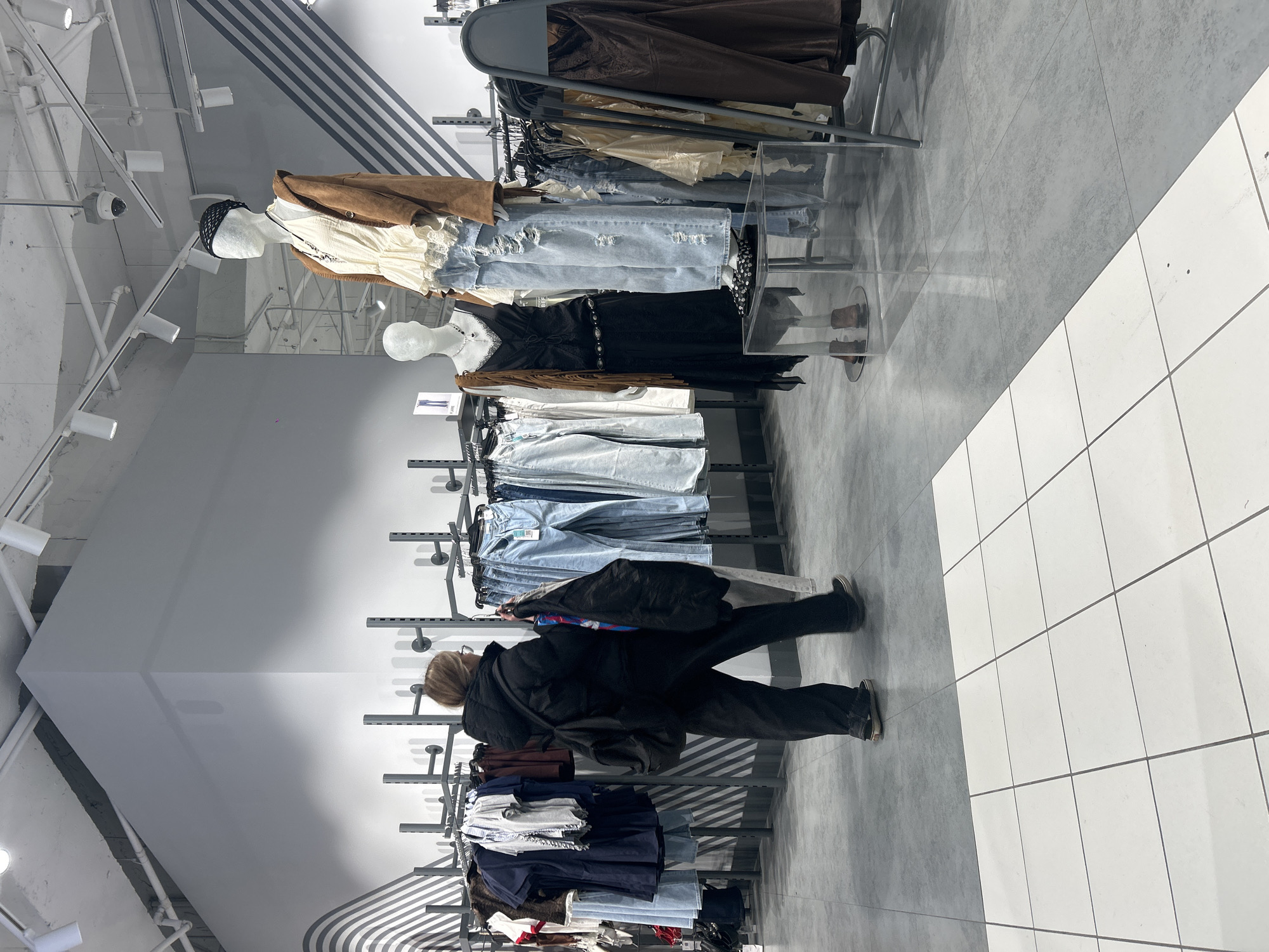 The image captures an indoor scene within a modern clothing retail store, likely an apparel section or a boutique. The perspective is from behind a customer, looking down an aisle.
**Subjects:** A person is visible from the back, appearing to be a female with light brown hair, wearing a dark, possibly padded, jacket and dark pants. She is moving alongside clothing racks. Several mannequins are prominently displayed, showcasing outfits. One mannequin wears a light brown jacket over a cream-colored top and light blue ripped jeans, with a black mesh head covering. Another mannequin further back is dressed in a black, patterned dress with a decorative belt. Numerous items of clothing, predominantly jeans in various washes (light blue, distressed, dark blue), as well as jackets and tops in neutral tones (brown, cream, black), hang on metal racks. The store's interior features white walls, some sections with grey and white diagonal stripes, a light grey ceiling with exposed white piping, and track lighting. A security camera is mounted on the ceiling. The floor is tiled, featuring a diagonal line that separates a grey tiled area from a white tiled area.
**Activity:** The person is actively browsing or walking through the clothing section of the store, presumably shopping.
**Notable details:** The environment is brightly lit by artificial ceiling lights, indicating an indoor setting. The general aesthetic is clean and modern. Small white tags or cards are visible on some clothing racks and near the mannequins, likely displaying product information or prices, though the text on them is illegible due to distance and resolution. No specific time of day or weather can be determined. The provided context indicates the location is Saint Petersburg, Russia.
**Visible text:** Small, illegible text appears on white tags or cards affixed to the clothing displays.