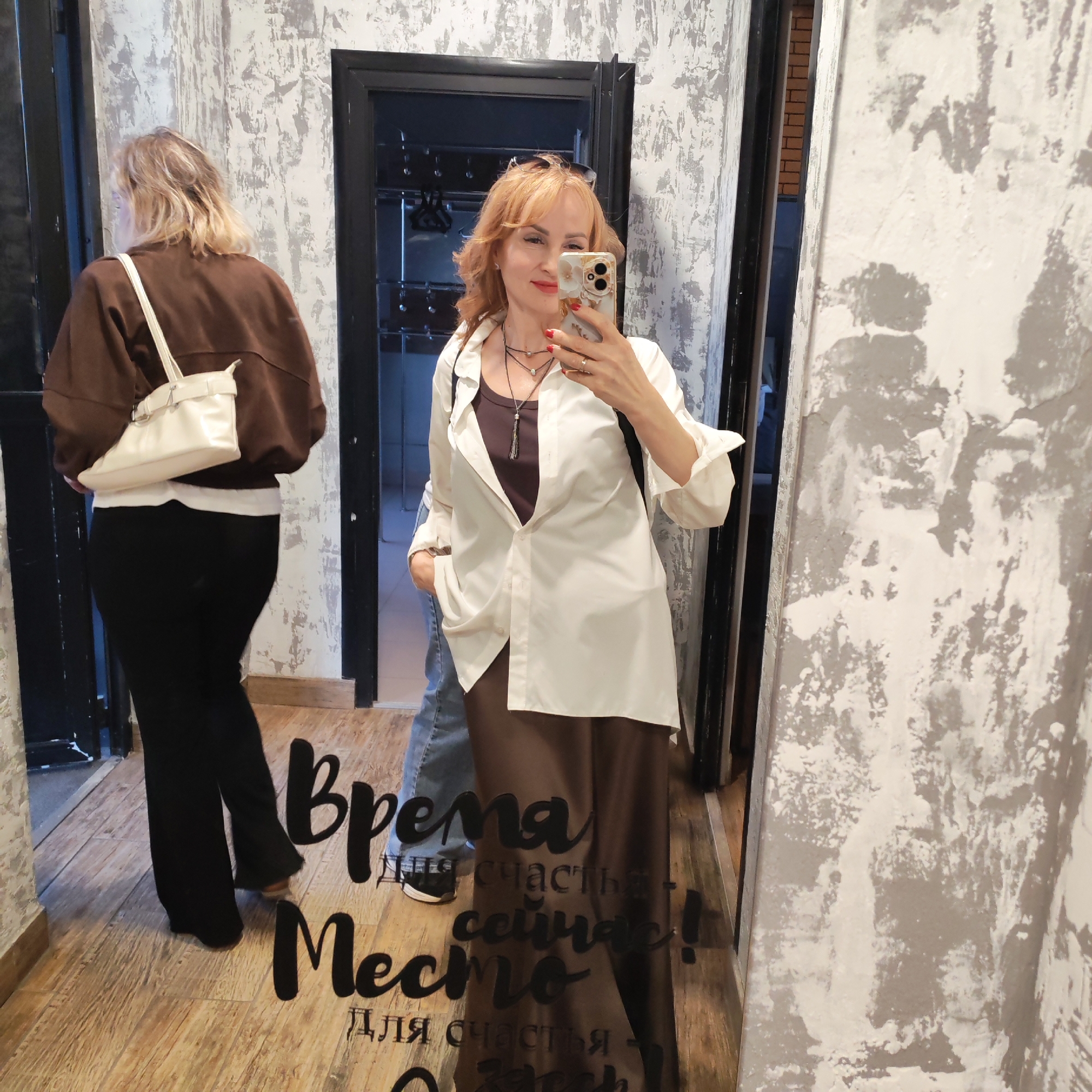 The image is a mirrored selfie taken in what appears to be a hallway or foyer of a building, potentially in Chaikovsky, Russia. The mirrored surface has the words "Время для счастья сейчас! Место для счастья здесь!" written on it.
The main subject of the image is a woman holding a smartphone up to take the photo. She has reddish-blonde hair, fair skin, and is wearing a brown top, a white button-down shirt, and a long brown skirt. She is wearing necklaces.
In the background, there is a second woman with blonde hair wearing a brown jacket, a white shirt underneath, and black pants. She is walking into or out of a doorway. A glimpse of another person wearing jeans can be seen behind the main subject.
The setting is a room with textured grey and white walls and wooden floors. There are doorways with black frames visible in the reflection.