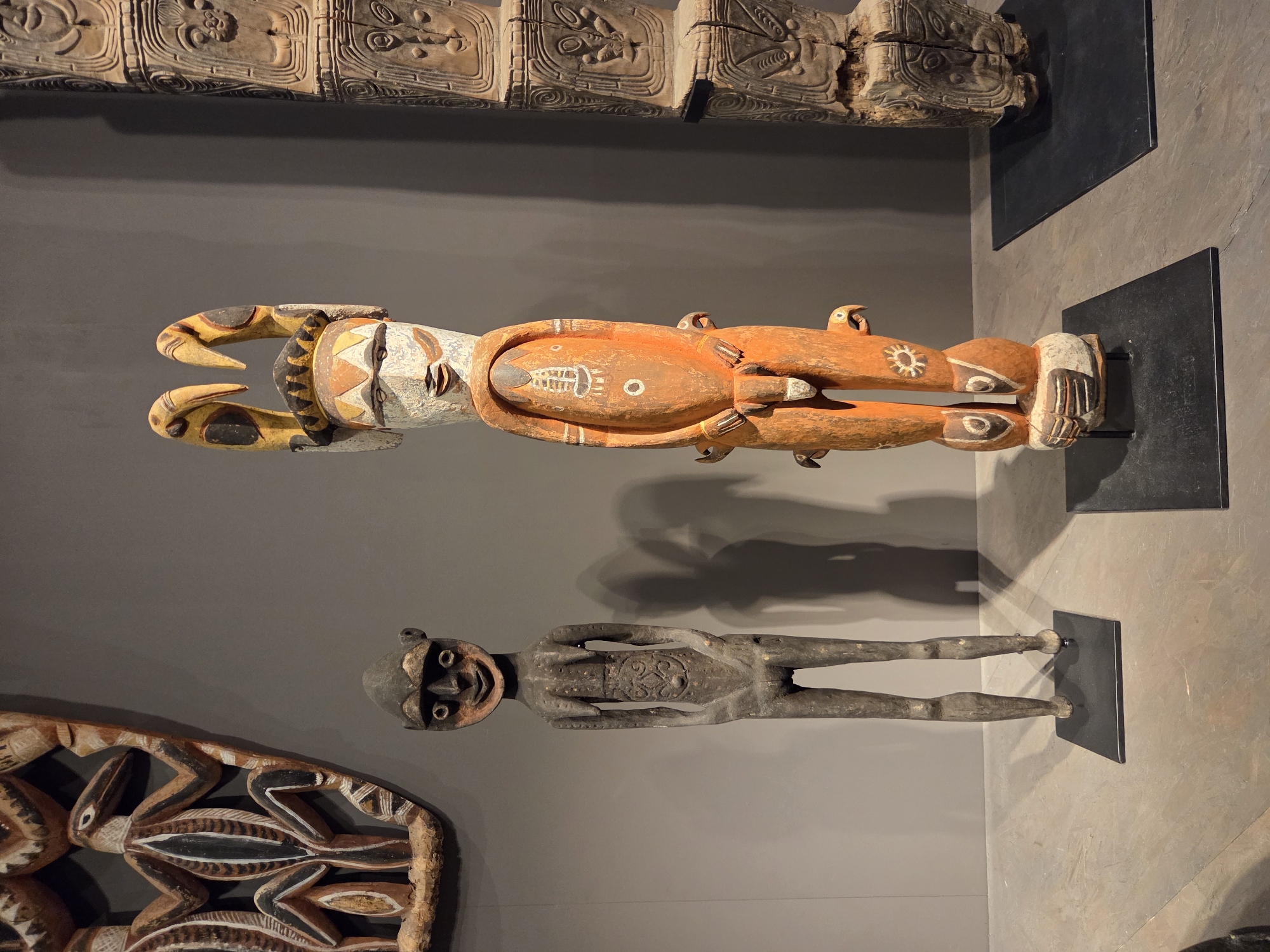 The image displays a collection of indigenous artifacts presented on a plain grey background in what appears to be a museum or gallery setting. In the foreground, two wooden sculptures stand on black display bases. The sculpture on the left is a dark, slender figure with a simplified facial structure and stylized body details, standing upright with hands at its sides. To its right, a larger, more colorful sculpture is depicted. This figure features a white, face-like mask with painted designs, a dark headdress with yellow horn-like protrusions, and an orange body with painted symbols. It is adorned with smaller bird-like figures attached to its sides and base. To the far left, a portion of a carved wooden mask or shield with intricate patterns and depictions of bird-like creatures is visible, mounted on the wall. On the far right, another carved wooden artifact, appearing to be a totem or column, stands vertically, displaying multiple carved faces and geometric patterns. The floor is a textured concrete or stone surface. The lighting casts shadows of the sculptures onto the wall behind them. The environment suggests an indoor exhibition space in Santa Ana, United States of America.
