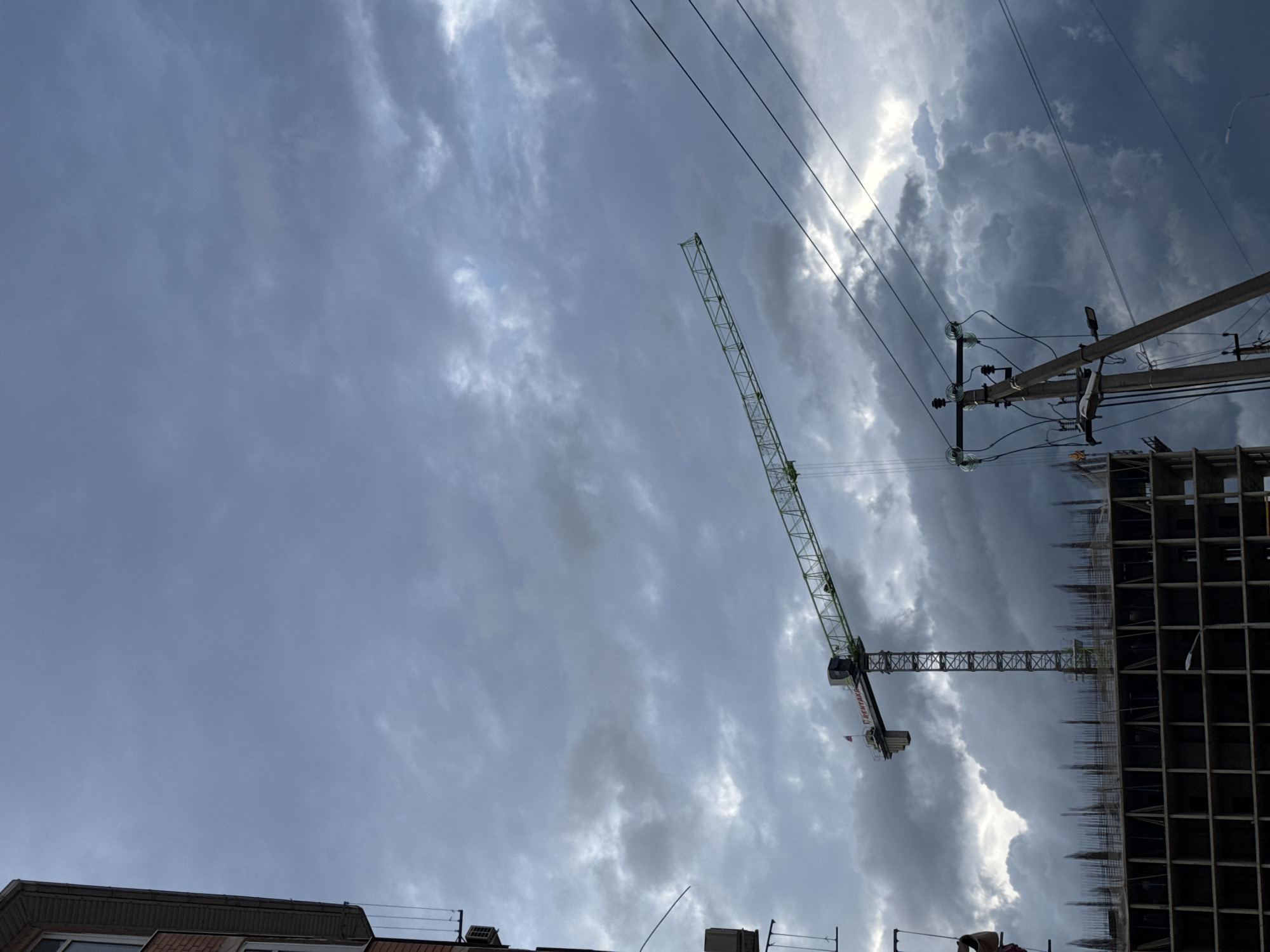 The scene captures a cloudy sky over an urban construction site in Unknown, Unknown. A tall green construction crane dominates the view, positioned next to a building under construction, its skeletal frame visible. Power lines run diagonally across the upper right of the frame, supported by a wooden utility pole with insulators. Building rooftops are visible along the bottom of the image.