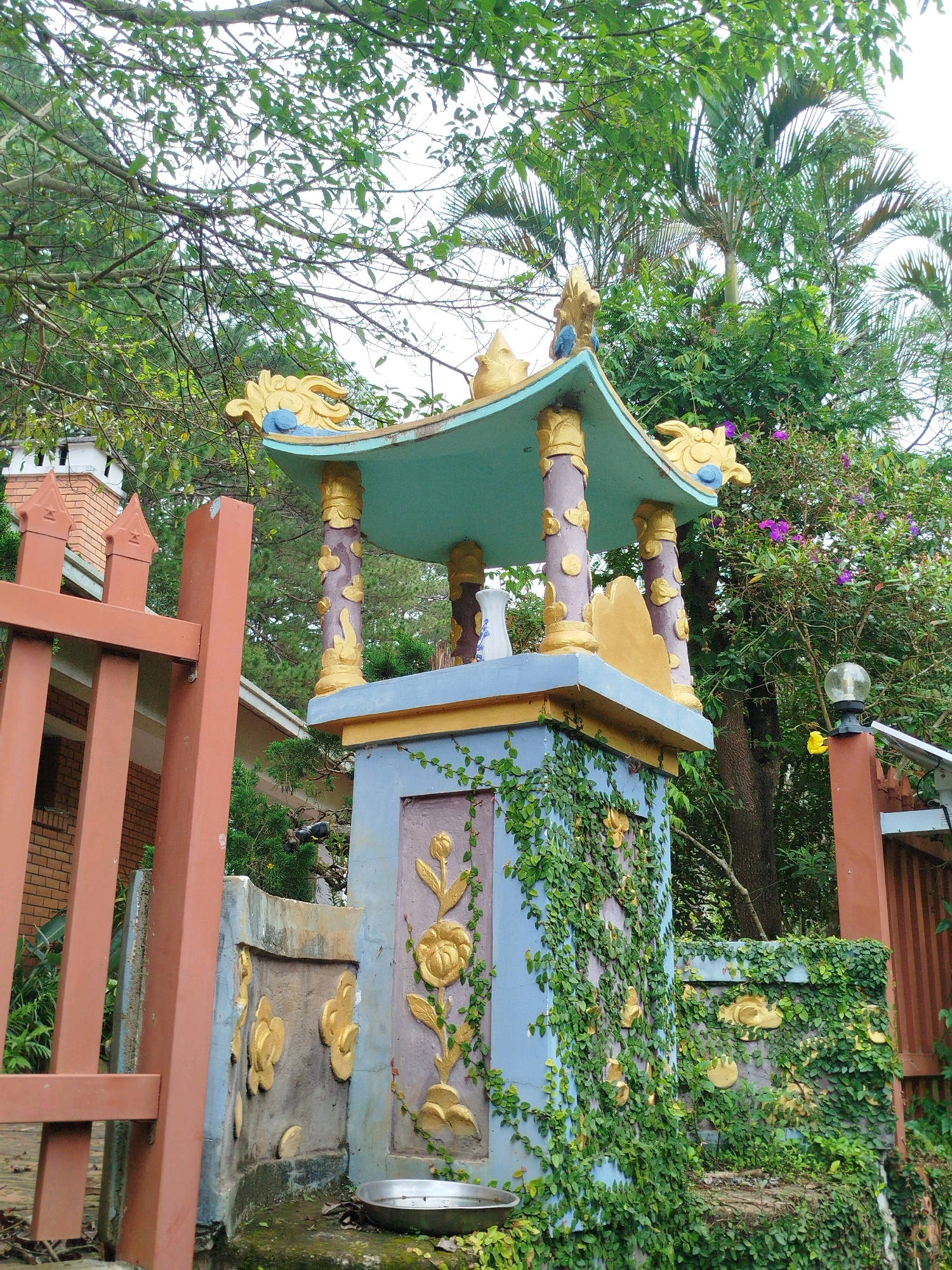 This is a close-up, eye-level, outdoor, daylight photograph of a shrine structure located in Da Lat, Vietnam. The shrine is ornate, with a teal-colored, curved roof adorned with gold dragon motifs and blue accents. It is supported by four pillars, which are painted in a purple hue with gold floral embellishments. The base of the shrine is a blue rectangular structure decorated with gold floral patterns, and it is heavily overgrown with green ivy. A silver metal bowl sits on the ground at the base of the shrine.
To the left of the shrine, a portion of a reddish-brown metal fence is visible, with a brick building behind it. To the right of the shrine, a reddish-brown fence post supports a black outdoor light fixture with a globe-shaped glass shade and a small solar panel. Lush green trees and foliage, including palm trees and flowering bushes with purple blossoms, surround the shrine, creating a natural and somewhat overgrown environment. The sky is overcast and visible through the tree canopy. No people are present in the image.