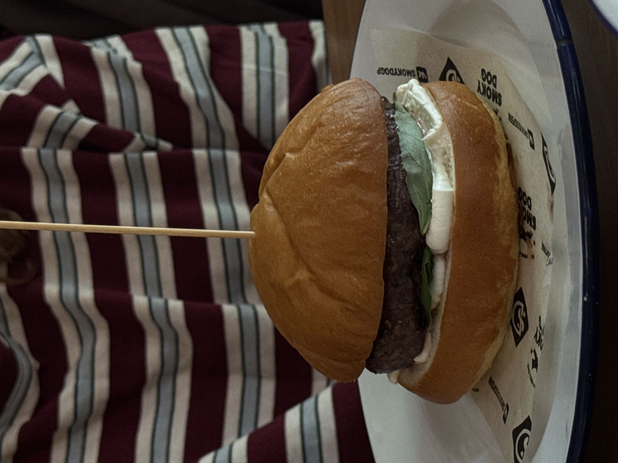 The image shows a burger, presumably served at a restaurant or food establishment called "Smoky Dog", as the logo on the paper wrapping indicates. The burger has a brown bun, a meat patty, greens, and a white sauce or cheese. A wooden skewer pierces the burger, likely to hold it together. The burger sits on a white plate with a blue rim. The background features a striped fabric with maroon, white, and light blue stripes. The image does not provide enough information to confirm the location in Perm, Russia.
