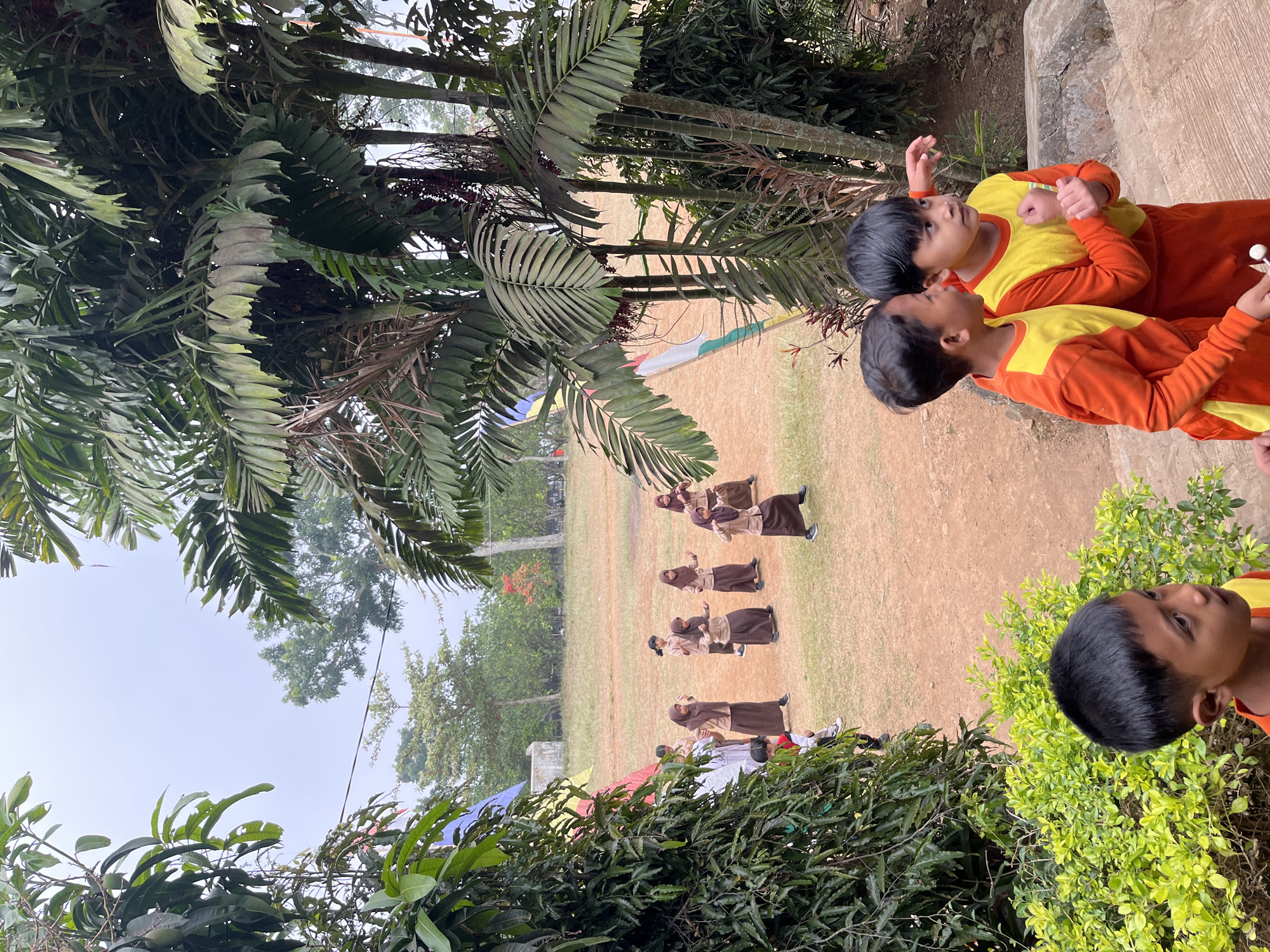 In Gembong, Indonesia, an outdoor event is underway on a grassy field with a dirt perimeter. The scene is partially framed by lush green foliage and palm trees. In the foreground, two young boys in vibrant orange and yellow uniforms stand facing away from the camera, looking towards the field. Behind them, another young boy in similar attire is partially visible. On the field, a group of children, dressed in brown hijabs and long brown dresses, are engaged in what appears to be a dance or performance. Some are in motion, while others are observing. A colorful banner, predominantly yellow, red, and blue, is visible to the left of the performing children. In the background, trees and a pale sky suggest an overcast day. The overall environment is natural and suggests a school or community gathering.