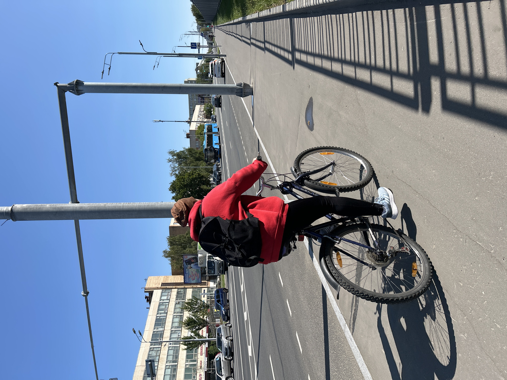 A person wearing a red hooded jacket and a brown beanie is riding a dark blue bicycle on a paved bike path. The person is carrying a black backpack. The bike path is adjacent to a multi-lane road with cars and a blue bus. In the background, there are several multi-story buildings and trees. The sky is clear and blue. The scene appears to be in a city, likely Moscow, Russia, given the presence of Cyrillic signage on the buildings and the overall urban infrastructure. The time of day is daytime, indicated by the bright sunlight casting shadows on the path.