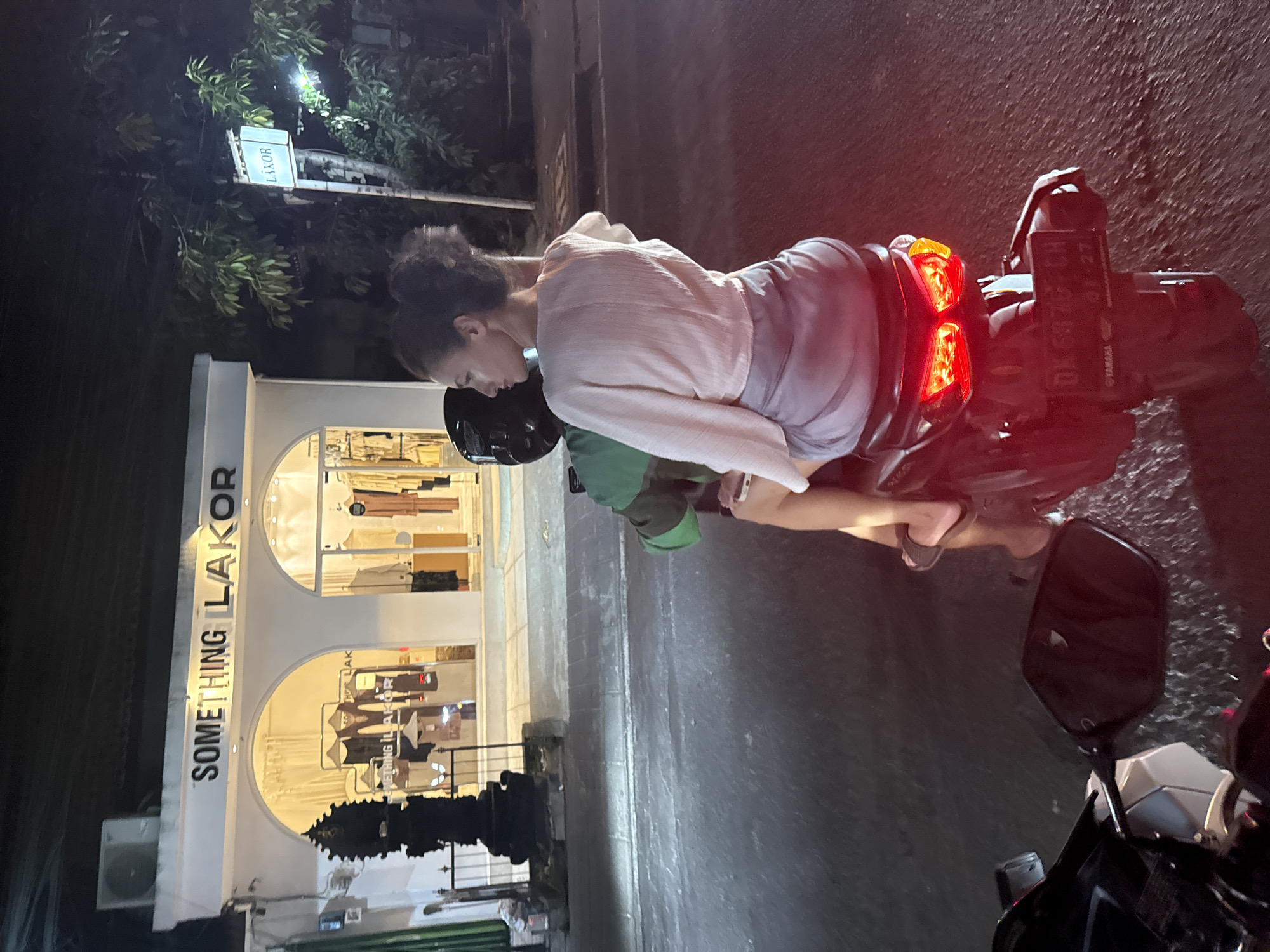 A woman is seen astride the back of a parked motorcycle at night on a street in North Kuta, Indonesia. The motorcycle's tail lights are illuminated and the license plate is partially obscured. The storefront of "Something Lakor" is visible behind her, with clothes displayed in the illuminated windows. Further up the street, a sign for "Lakor" is also illuminated above trees. The street is paved.