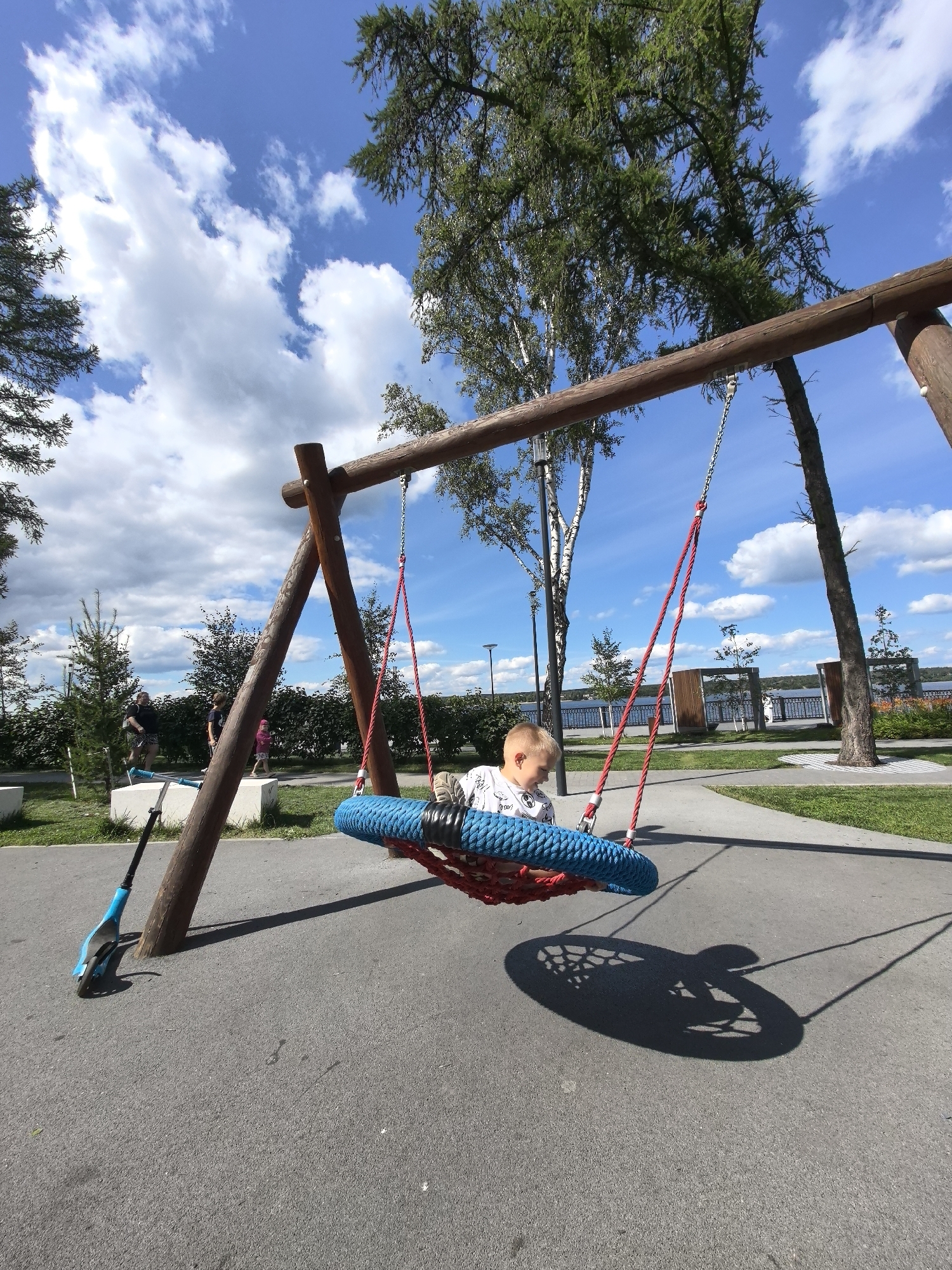A young boy sits on a large, circular blue net swing suspended from a wooden A-frame swing set. He is wearing a white t-shirt with a black graphic. The swing is casting a shadow on the asphalt playground surface. To the left of the swing set, a blue and black kick scooter is propped up against one of the wooden support beams. In the background, several trees with green foliage are visible under a bright blue sky with scattered white clouds. Other people, including a man and a child, are visible in the middle distance, walking on a paved path. The playground appears to be located in a park area with green grass and a body of water, likely a river or lake, visible beyond a decorative fence in the far background, suggesting a location near the Kama River in Perm, Russia.