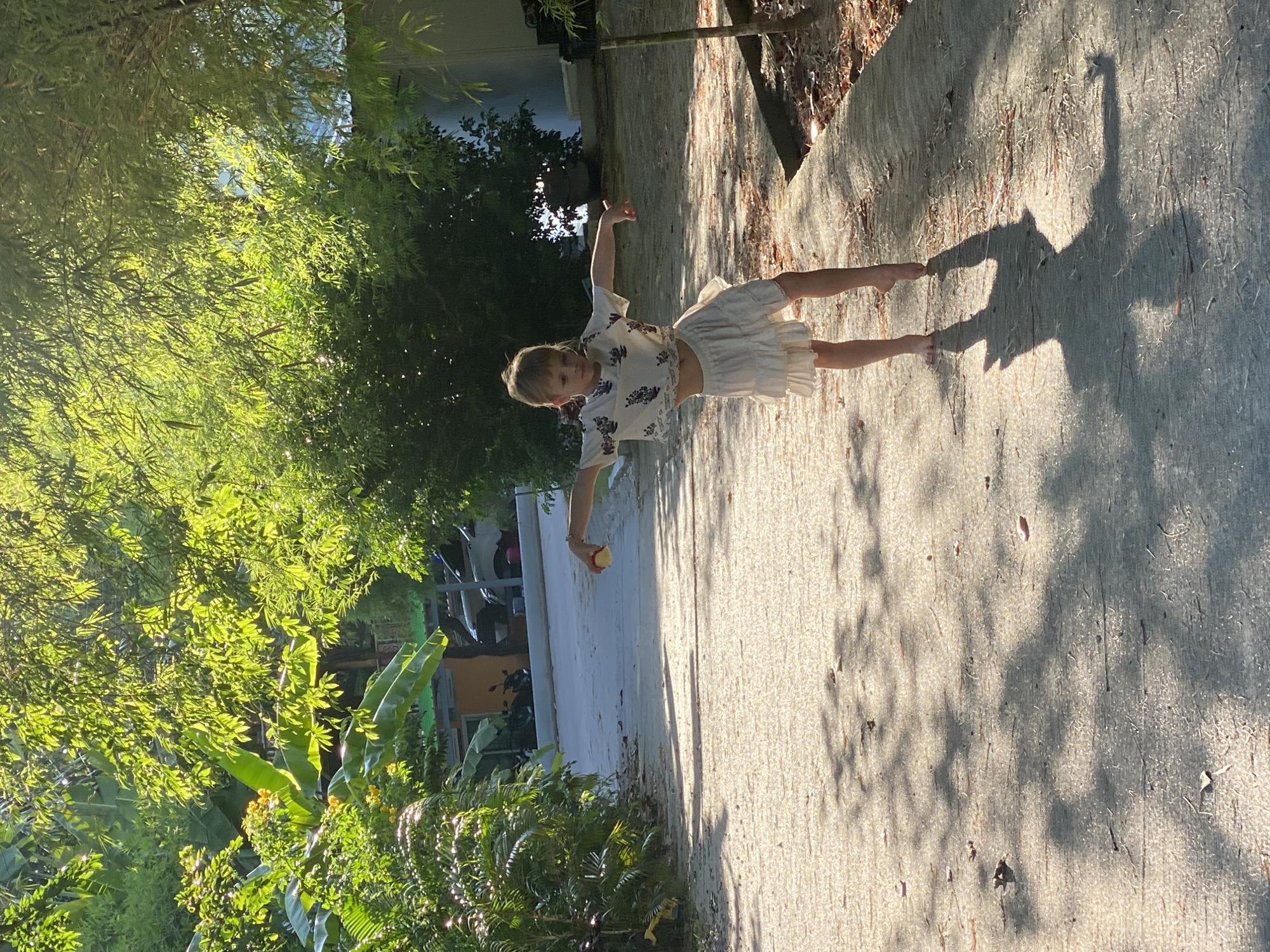 A young child, appearing to be female with blonde hair, is captured mid-stride on a paved pathway bathed in dappled sunlight. The child is wearing a white, cropped t-shirt with a floral pattern and a tiered, white ruffled skirt. Their arms are outstretched, one slightly higher than the other, in a pose that suggests dancing or playful movement. The child is barefoot. The pathway is surrounded by lush green vegetation, including broad-leafed plants and bamboo. Sunlight filters through the leaves, casting distinct shadows on the ground. In the background, a building with a yellow facade and a gray roof is visible, along with parked vehicles including motorcycles and a car. The overall environment suggests a tropical or subtropical setting, possibly a residential area or a pathway through a garden or natural park. The location is identified as Ko Pha Ngan, Thailand, specifically in the Tambon Ko Pha Ngan.