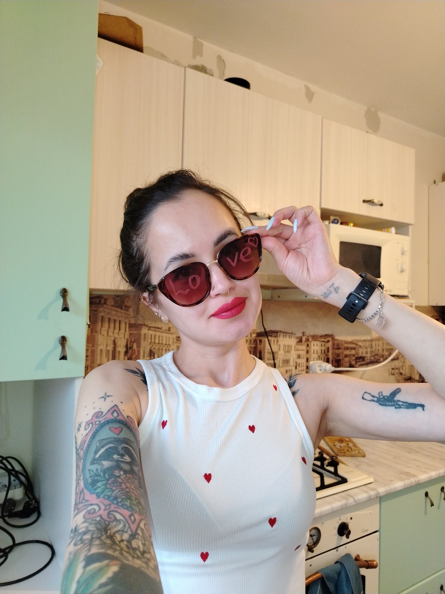 A woman is taking a selfie in a kitchen. She is wearing sunglasses with the word "love" on one lens and a white tank top with small red hearts. She has tattoos on both arms. The kitchen has light green and white cabinets, a microwave, and a stove. The background features a mural of a European city. The city depicted in the mural is not Noviy, Russia.