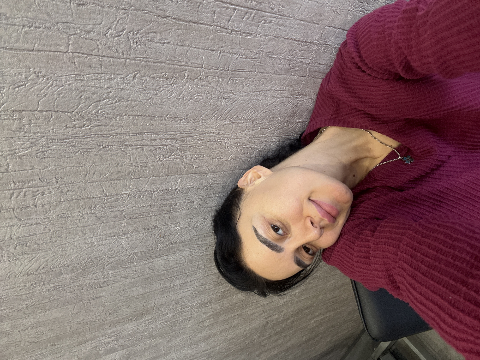 An adult female is depicted in a close-up selfie, oriented slightly diagonally within the frame. She possesses dark hair, pulled back, revealing her ears. Her eyebrows are dark and prominent, framing light brown eyes. A slight smile is present on her lips, and her complexion appears natural with minimal makeup. She wears a deep magenta or plum-colored, V-neck, long-sleeved, ribbed knit sweater. A silver-toned necklace with a small, light blue or green cross-like pendant hangs around her neck. Her head is tilted, with her left cheek resting gently against her left shoulder, which is partially visible. The background consists of a textured wall, colored in a muted gray-brown or taupe, featuring vertical, irregular striations and raised patterns resembling rough plaster or bark. A small segment of a dark, possibly black, chair back is visible in the lower-left corner of the frame. The image does not contain any visual identifiers to specify a particular location within the city of Tbilisi, Georgia.