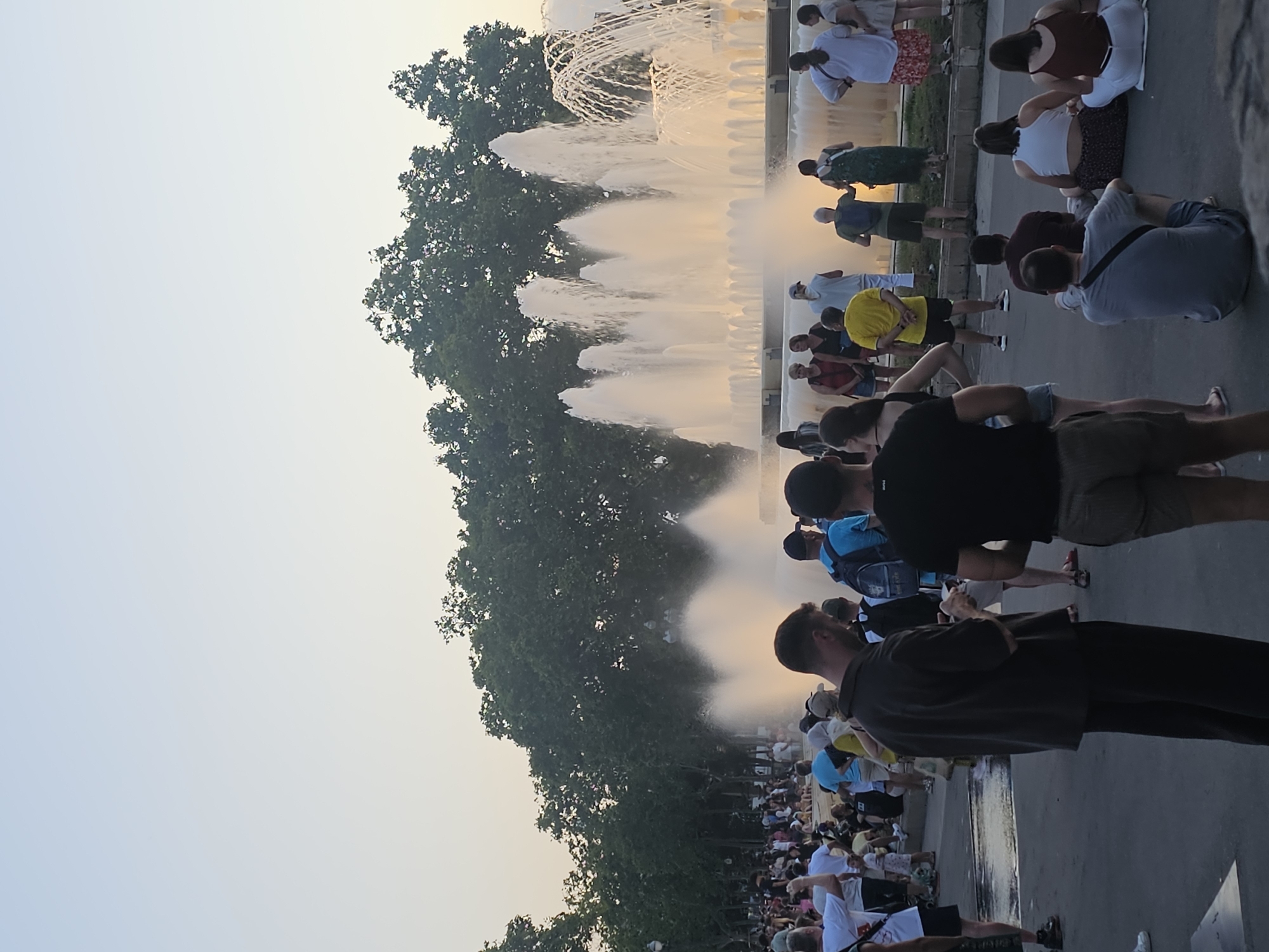 The image shows a crowd of people gathered near a large, multi-tiered fountain. The fountain is spraying water high into the air, creating a misty, dynamic spectacle. The setting appears to be an outdoor public space, possibly a park or plaza in Barcelona, Spain, given the context.
The people in the image are of various ages and are dressed in casual summer clothing, suggesting a warm, likely late afternoon or early evening time of day, as indicated by the soft, warm light casting long shadows. Many individuals are standing, watching the fountain, while others appear to be sitting or walking along the edge of the fountain or a pathway. Their postures and the general gathering suggest a leisurely atmosphere, with people enjoying the visual display.
Notable details include the lush green trees bordering the fountain on the left side of the image, providing a natural contrast to the man-made structure. The sky is a pale, hazy blue, hinting at either a bright day with some atmospheric diffusion or perhaps early dusk. There are no visible signs or text within the frame that provide specific information about the location or event. The overall mood is one of shared enjoyment and relaxation, as people come together to appreciate the water feature.