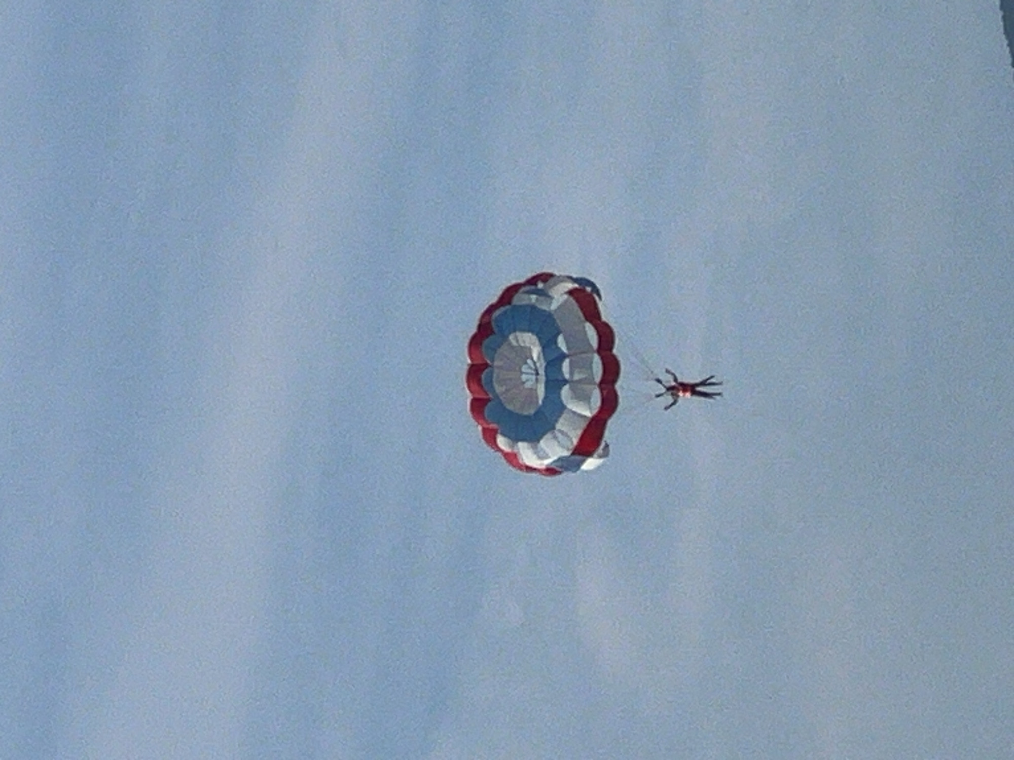 A paraglider, wearing a red suit, is suspended in the sky over Đà Nẵng, Vietnam, under a large, circular parachute. The parachute features a pattern of red, white, and blue sections. The sky is a clear blue with some light clouds.