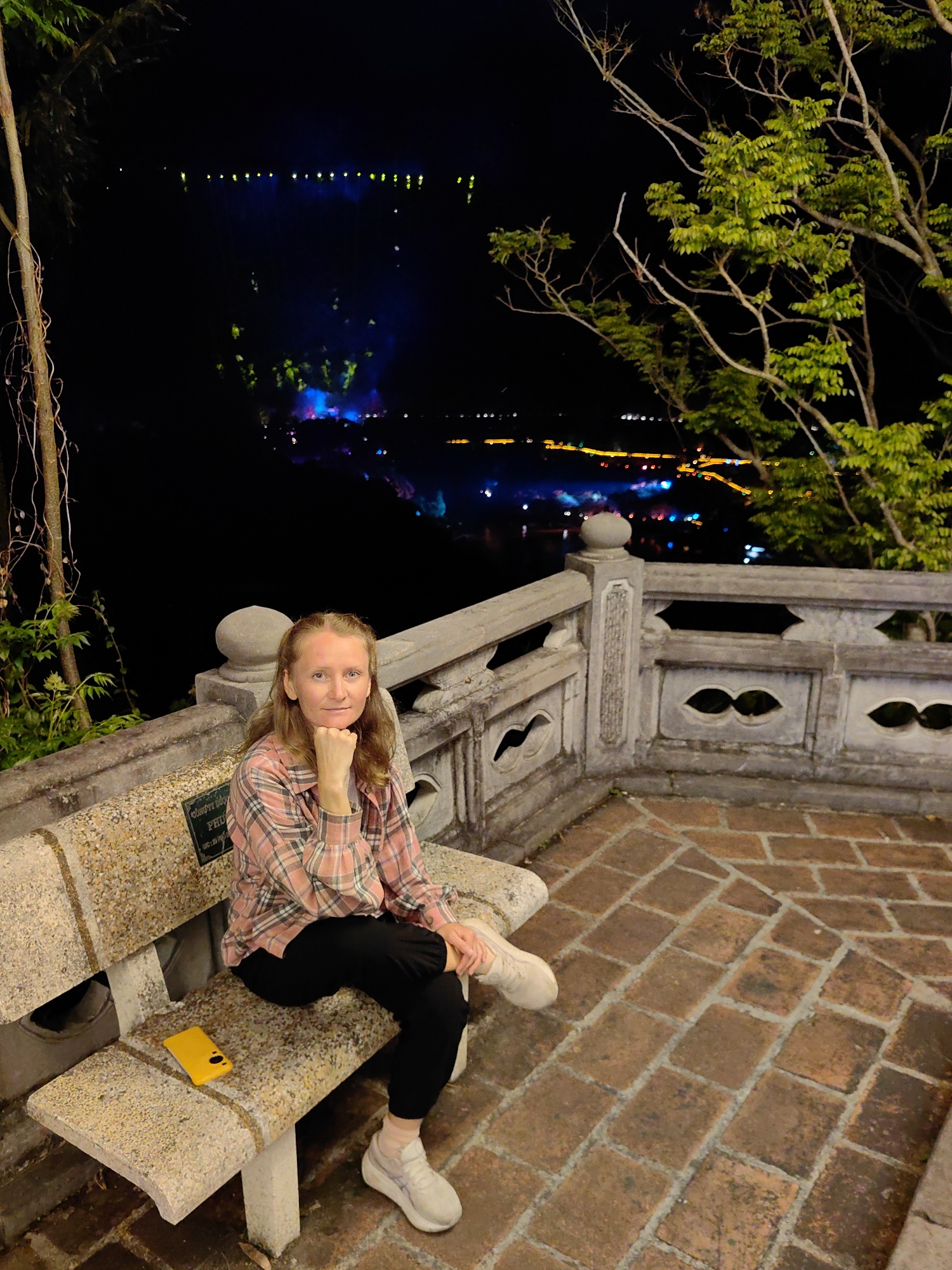 A woman is sitting on a stone bench in Xã Đàm Thủy, Vietnam, at night. The woman is wearing a pink and black plaid shirt, black pants, and light-colored sneakers. She has her right hand on her chin. A yellow cellphone rests on the bench to her left. The bench is part of a stone enclosure with decorative openings. The ground is paved with red brick. In the background is a dark landscape with lights scattered across the horizon and hints of structures illuminated with blue and green lights. Bare branches and leafy trees are visible on the right side of the frame.