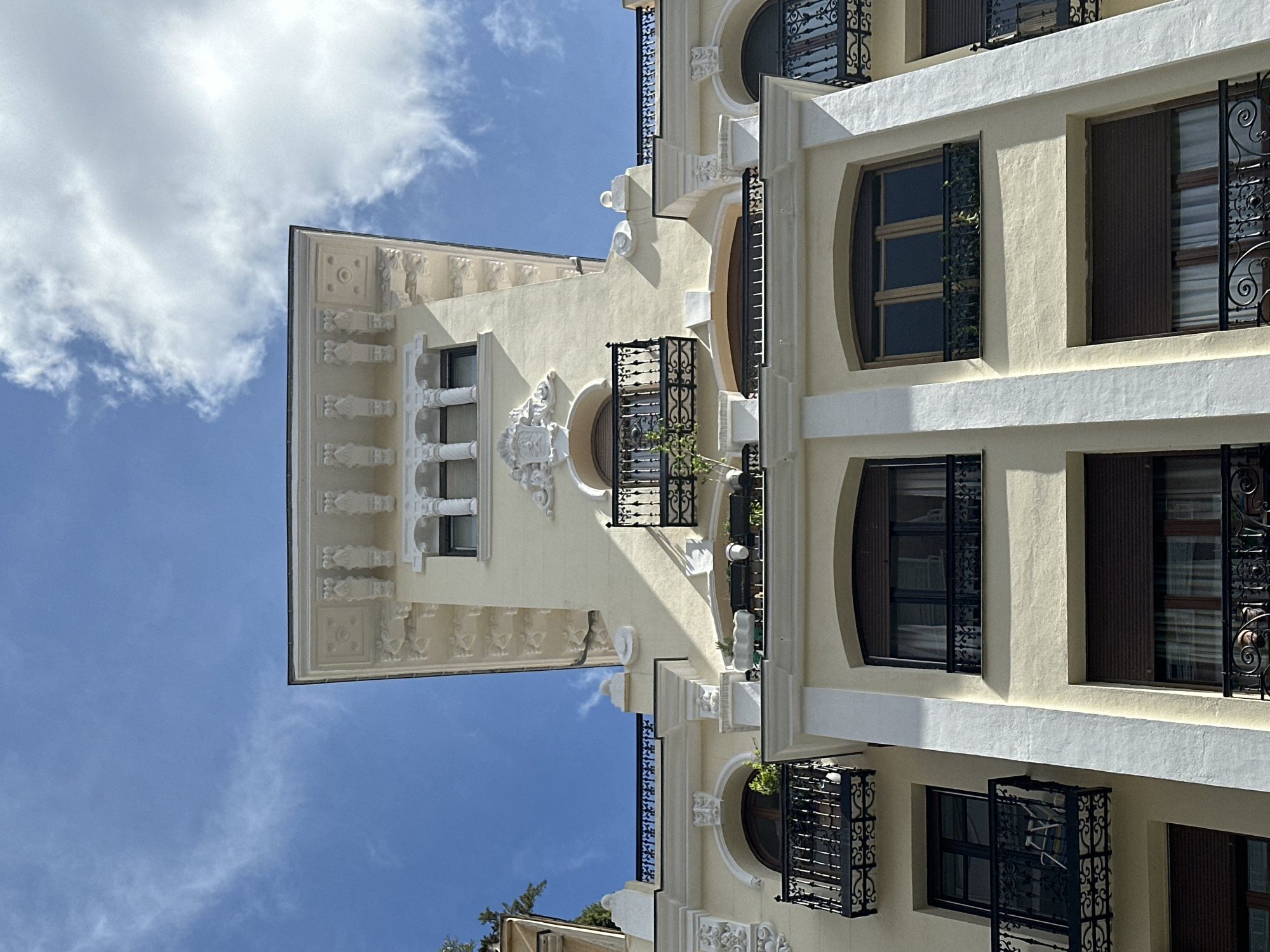 The image presents a view of an ornate building facade under a partly cloudy sky. The building is characterized by a pale yellow stucco exterior adorned with white trim. The architecture features round windows and balconies with black wrought iron railings. The facade shows intricate detailing and embellishments. Based on architectural style, this building is located in Madrid, Spain. There are no people visible.