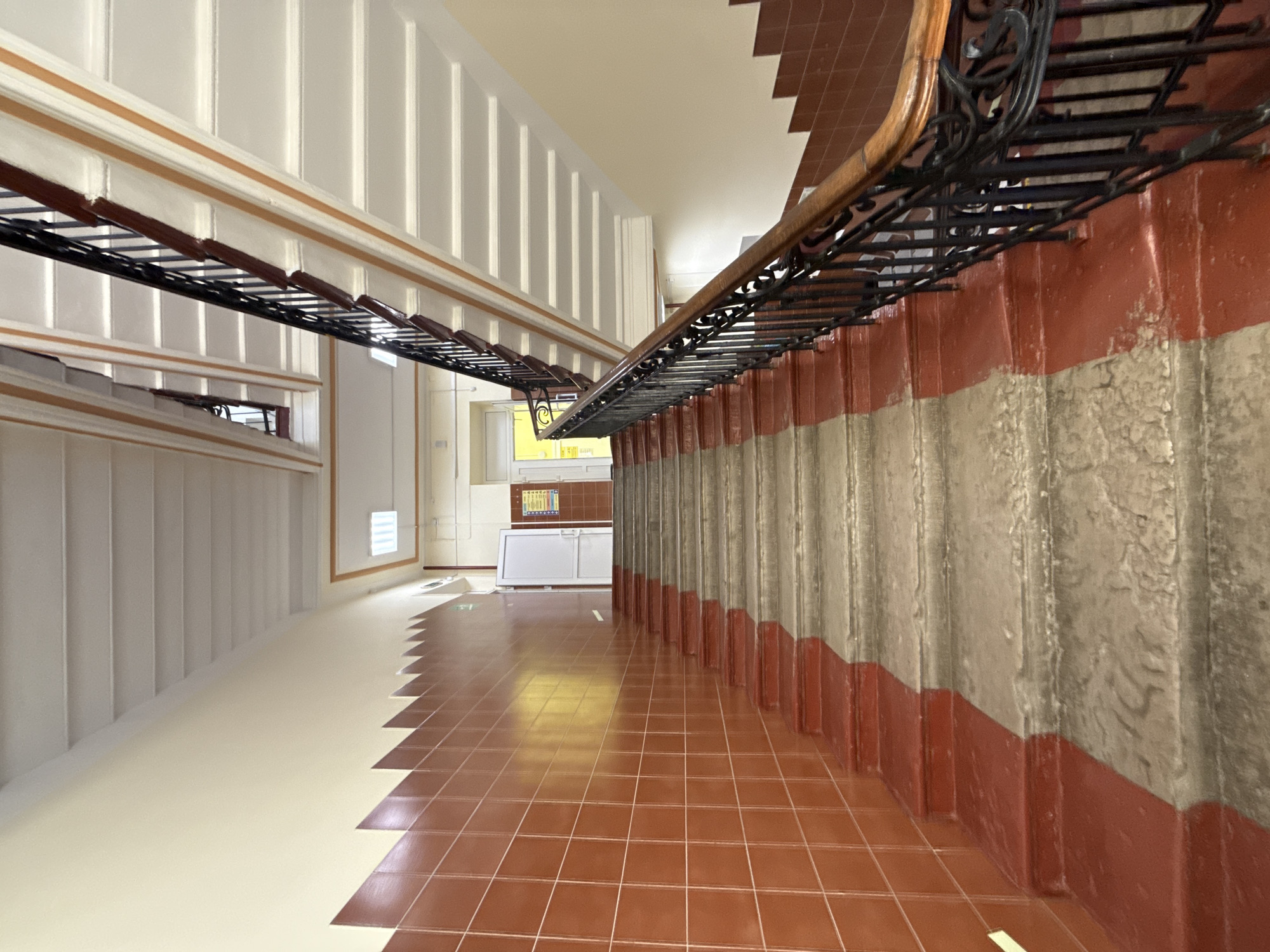 The image depicts a hallway with tiled floors and walls. The lower portion of one wall is red, and the upper portion is gray. The opposite wall is white with vertical rectangular indentations. A railing runs along the top of the red and gray wall. At the end of the hallway, there is a window and a counter. The image is taken in a hallway in Saint Petersburg, Russia.