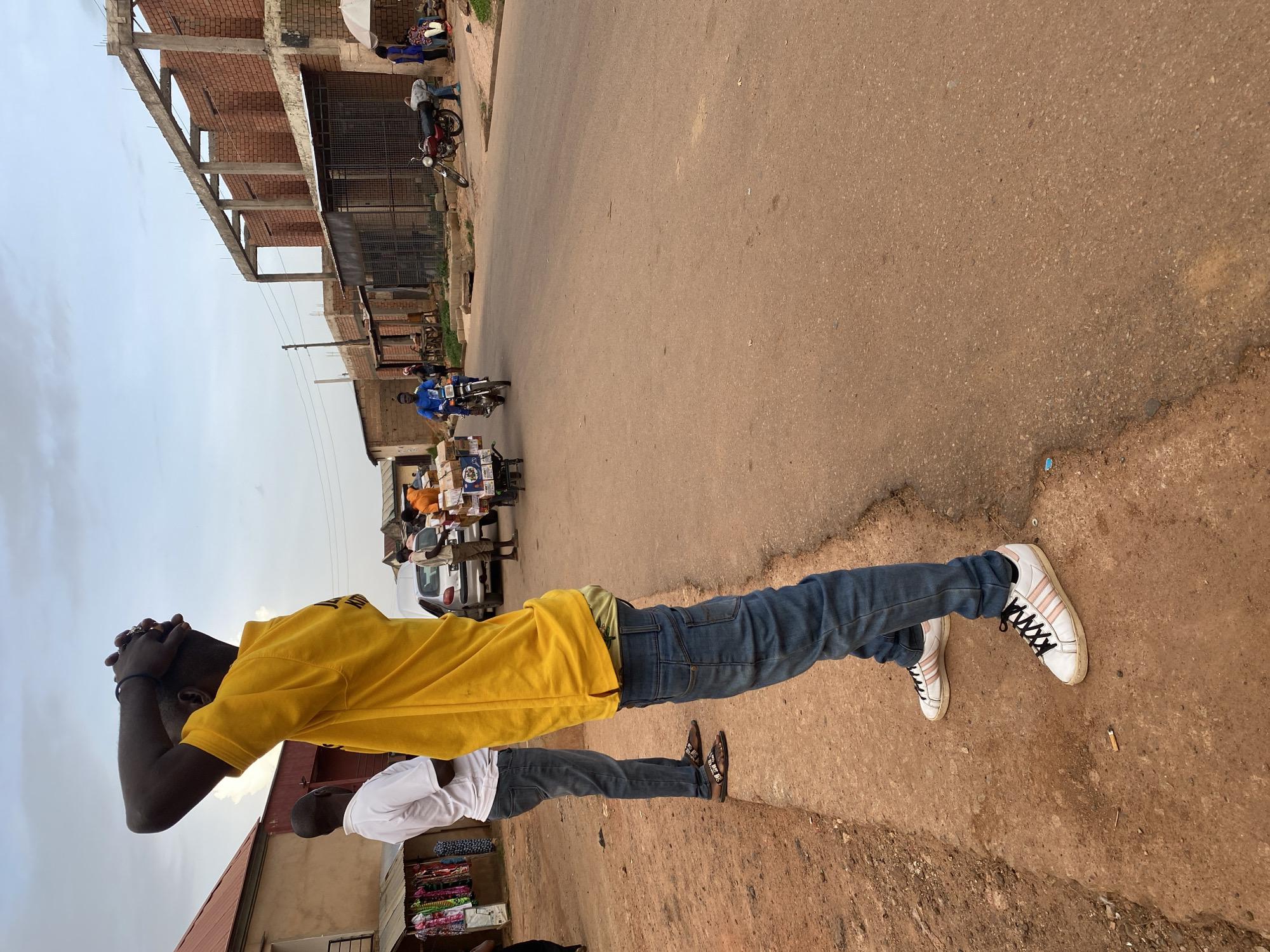 In Bukuru, Nigeria, this image captures a street scene. In the foreground, a Black man is seen from the waist down, facing away from the camera. He is wearing a bright yellow t-shirt, blue jeans, and white sneakers with pink and black stripes. His hands are clasped behind his head, and he appears to be looking up at the sky. To his left, another man in a white t-shirt and blue jeans stands with his arms by his side, also facing away from the camera.
The street is paved with reddish-brown asphalt. Several vehicles are present, including a motorcycle with a rider in a blue shirt, a silver car, and a red motorcycle parked on the side of the road. Other individuals are visible in the background, engaged in various activities. Buildings line the street, with one under construction made of red brick and concrete beams. In the distance, stalls with clothing are displayed. The sky is overcast, suggesting a cloudy day. The overall atmosphere suggests a typical day in a bustling urban environment.