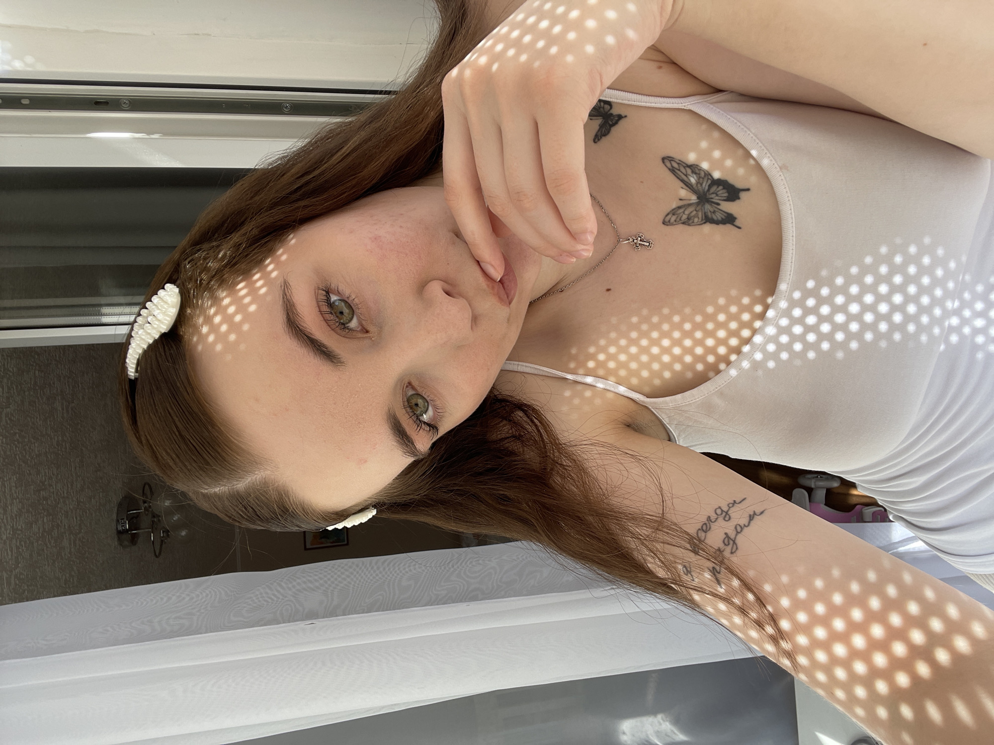 A fair-skinned woman is facing the camera. She has long brown hair and is wearing a white top. She has a cross pendant around her neck, butterfly tattoos on her chest, and other tattoos on her arm. The woman is in an interior setting, likely near a window with a curtain. The light filtering through the window creates dotted shadows on her face and chest. The scene is Chaikovsky, Russia.