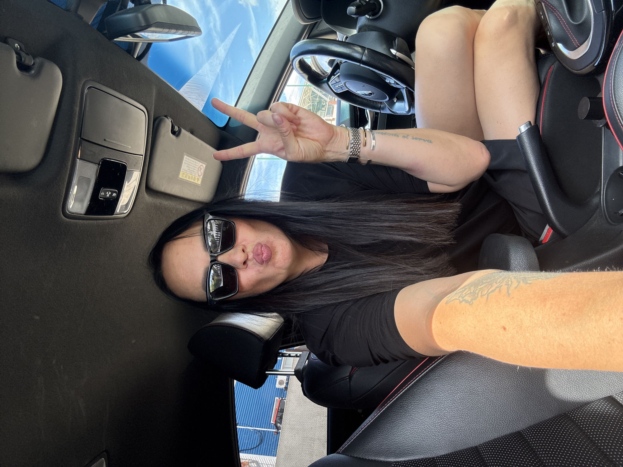 The image shows a woman sitting in the driver's seat of a car. She is wearing sunglasses and a black t-shirt, and she is making a peace sign with her right hand. The car's interior is visible, including the steering wheel, dashboard, and seats. The background shows a blue building with white trim, and a clear sky with some clouds. The location is identified as Chaikovsky, Russia.