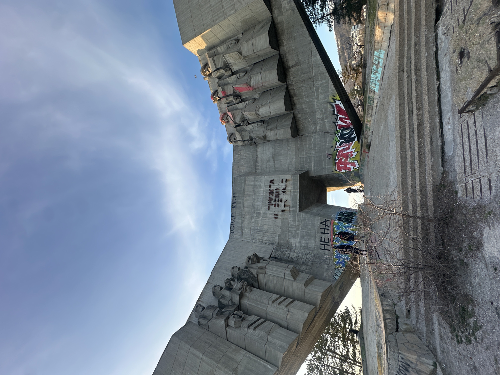 A low-angle shot captures a Brutalist-style concrete monument in Varna, Bulgaria, against a pale blue sky with wispy clouds. The monument features relief sculptures of figures, marred by graffiti in several places. Notably, "CALVIN KLEIN" is tagged in black and red above one set of sculptures. The Bulgarian phrase "НЕ НА" is stenciled in black on the side of the structure, next to colorful graffiti. Cracks are visible in the concrete, suggesting age or disrepair. A bare tree is in the foreground, partially obscuring the view. A person can be seen walking through an archway in the monument. Concrete steps lead towards the archway.