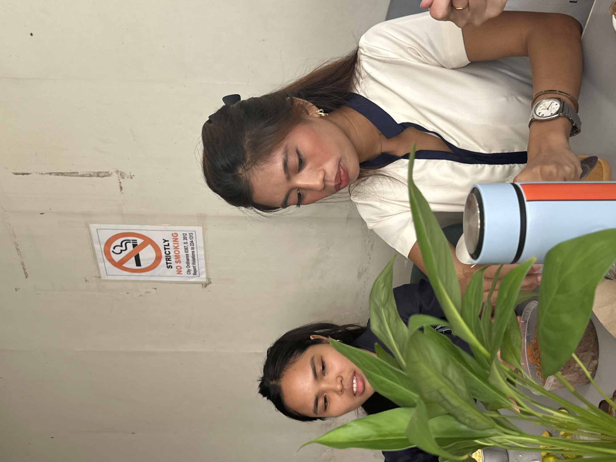 In Davao City, Philippines, two young women are seated at a table. One woman, on the left, has dark hair pulled back and is looking downwards with a slight smile. The other woman, on the right, has her dark hair tied up with a black clip and is looking down and to her left. She is wearing a white shirt with a navy blue collar and trim, a gold necklace, and gold hoop earrings. A silver wristwatch is visible on her left wrist, along with a bracelet. In the foreground, a large green plant with broad leaves partially obscures the view. On the table, a light blue and orange insulated flask is positioned to the right of the plant, and to the right of the flask, a person's hands are visible, one wearing a gold ring. A "STRICTLY NO SMOKING" sign is affixed to the light-colored, textured wall behind the women. The environment appears to be an indoor dining or seating area.