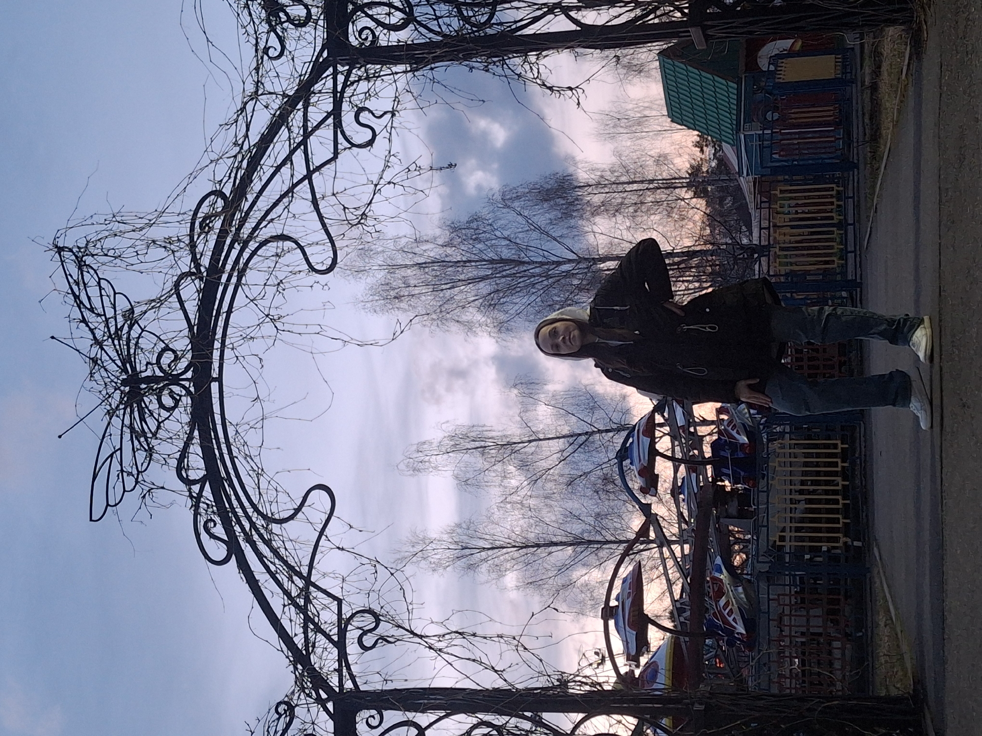 The image depicts an outdoor scene featuring a young person standing under an ornate metal arch. The arch, dark in color, is adorned with intricate scrollwork and covered in numerous bare, leafless vines or branches, suggesting a colder season. The person, who appears to be female, is seen from the mid-thigh up, wearing a dark winter jacket, blue jeans, and light-colored sneakers. She has a light-colored hood or hat on and is looking directly at the camera with a neutral to slight smile.
In the background, there is a children's playground area. Visible elements include a structure with a bright green peaked roof and what appears to be a small carousel or ride with colorful, boat-like seats. Beyond the playground, several deciduous trees with bare branches are silhouetted against the sky.
The sky dominates the upper portion of the scene, displaying a soft gradient of colors, ranging from light blue to shades of gray, pink, and orange. This coloration suggests either early morning (sunrise) or late afternoon (sunset), with diffused light indicating some cloud cover. The overall atmosphere is calm and quiet, characteristic of a park or recreational area in a temperate or cold climate, consistent with the location context of Chaikovsky, Russia. No visible text is present in the image.