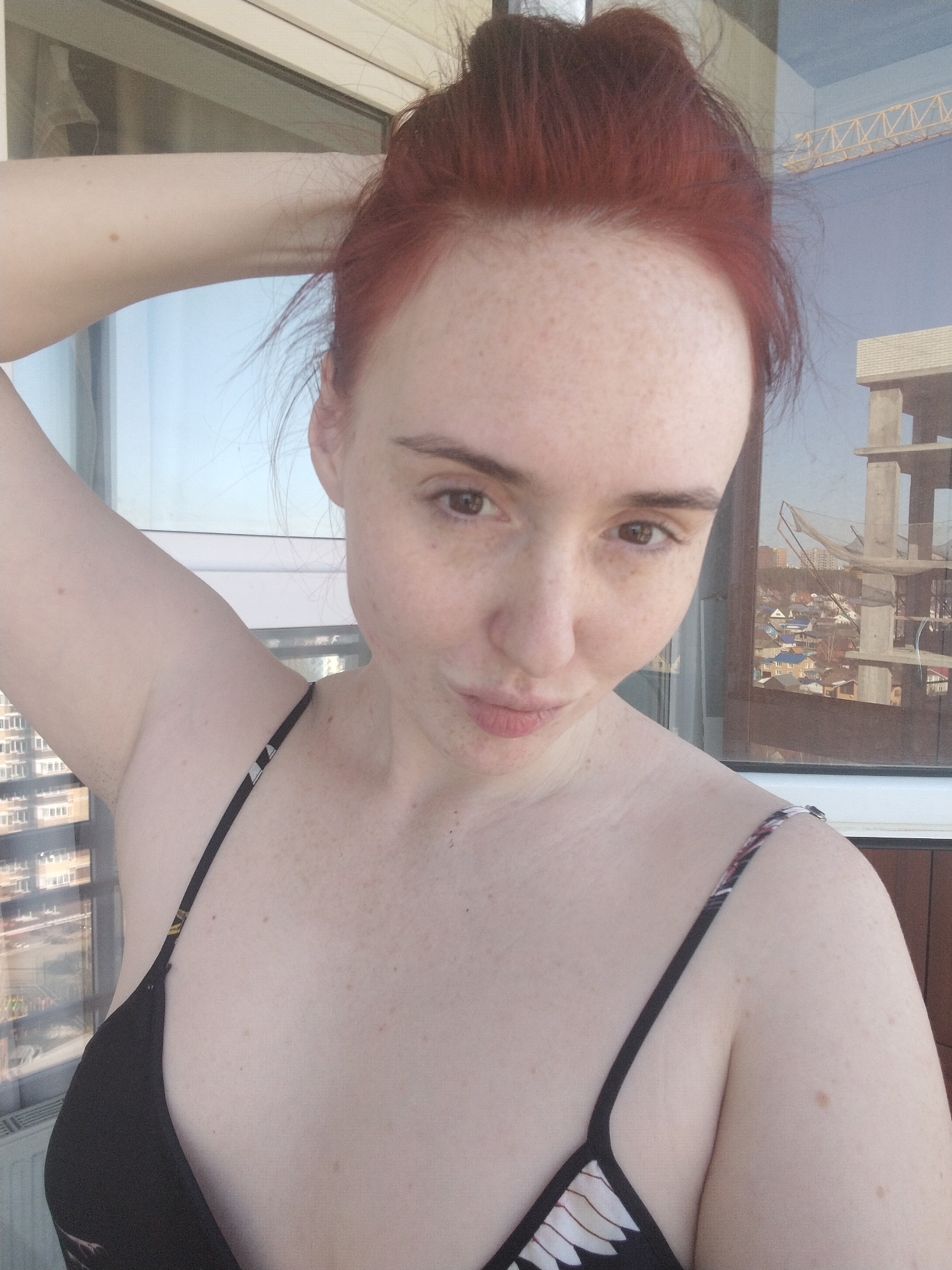 A selfie shows a fair-skinned, red-haired woman with freckles, wearing a black top with patterned straps. Her hair is in a messy bun. She has one arm raised behind her head. The background consists of a window reflecting urban scenery, including a construction site with a crane and partially constructed buildings, along with other city buildings and rooftops. The photo appears to be taken in an indoor location. Based on visual characteristics, the location appears to be Izhevsk, Russia, identifiable by the distinct architecture.