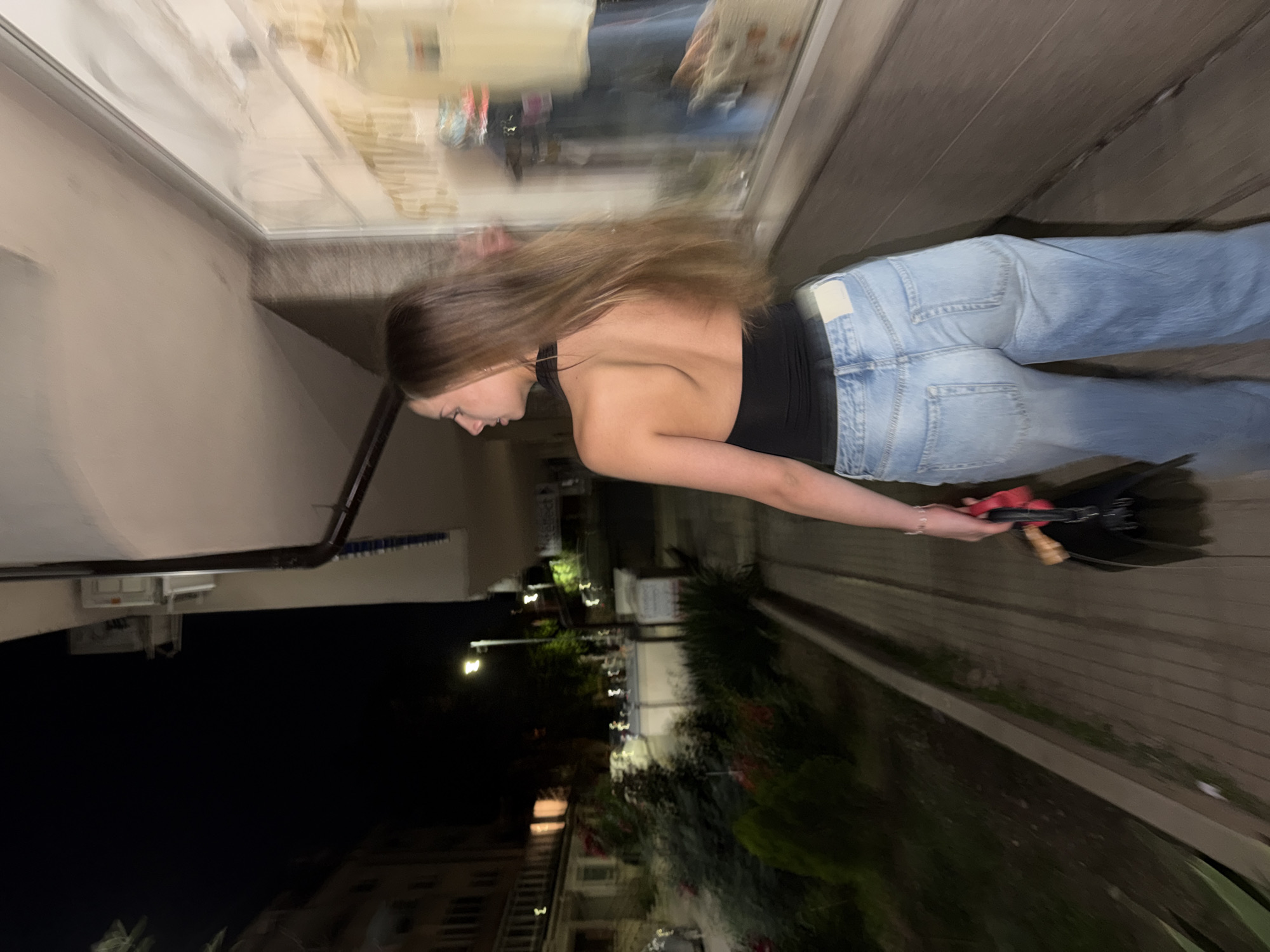 A young woman with long, light brown hair is captured from behind, walking away from the camera at night. She is wearing a black halter top and light-wash, wide-leg jeans. She is holding a black bag with red accents in her right hand and a delicate silver bracelet adorns her left wrist. The scene appears to be on a walkway next to a building with a storefront window, possibly in Sofia, Bulgaria, given the context. The window displays clothing items, and the street beyond is dimly lit with streetlights and lights from other buildings. The overall impression is a casual nighttime stroll in an urban environment.