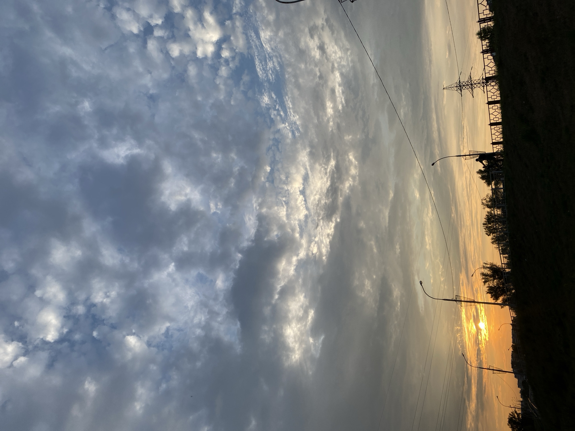 The image depicts a sunset scene in Minusinsk, Russia. The sky is filled with a mix of dark and light clouds, with the sun peeking through the clouds on the left side of the frame, casting an orange glow. In the foreground, a dark silhouette of a grassy embankment is visible. On top of the embankment, a decorative fence runs horizontally across the middle of the image. Behind the fence, silhouetted trees and buildings are visible. Several streetlights with wire baskets are positioned along the embankment. In the middle ground, there are power lines crisscrossing the sky. On the right side, a tall power pylon stands against the sky. A person on a bicycle is silhouetted against the sky, riding along the embankment.